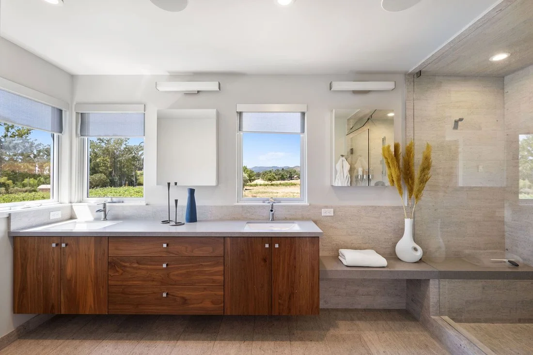 Modern bathroom with wooden vanity, dual sinks, gold-colored decor, large windows with blue shades, and a walk-in shower with glass door.