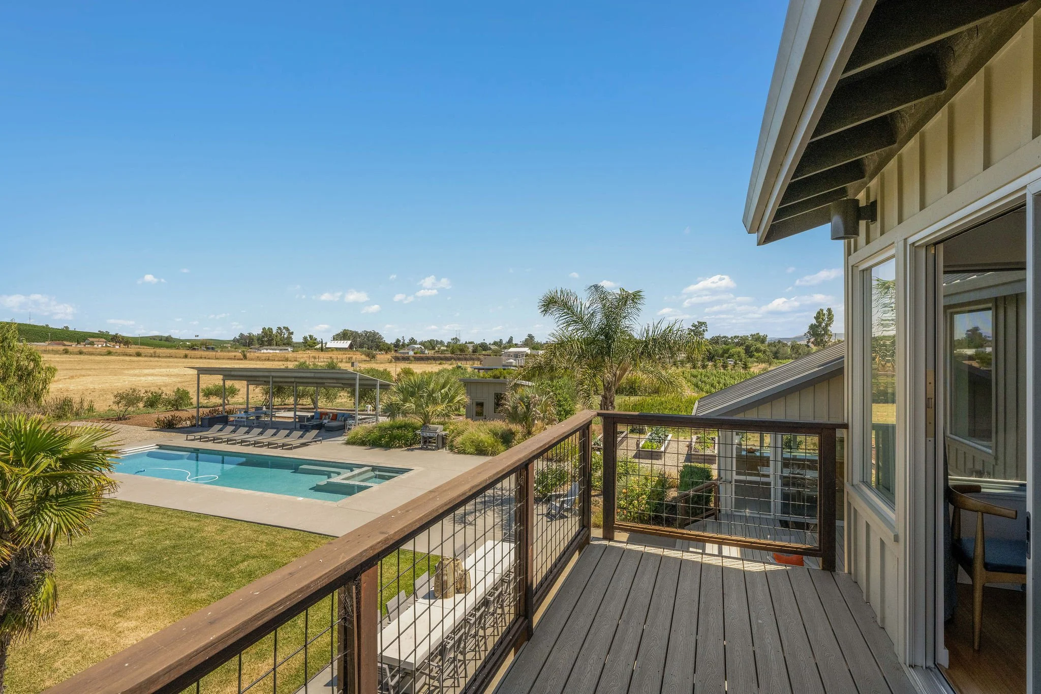 View from a balcony overlooking a backyard with a swimming pool, lounge chairs, and lush greenery, with open fields and a blue sky in the background.