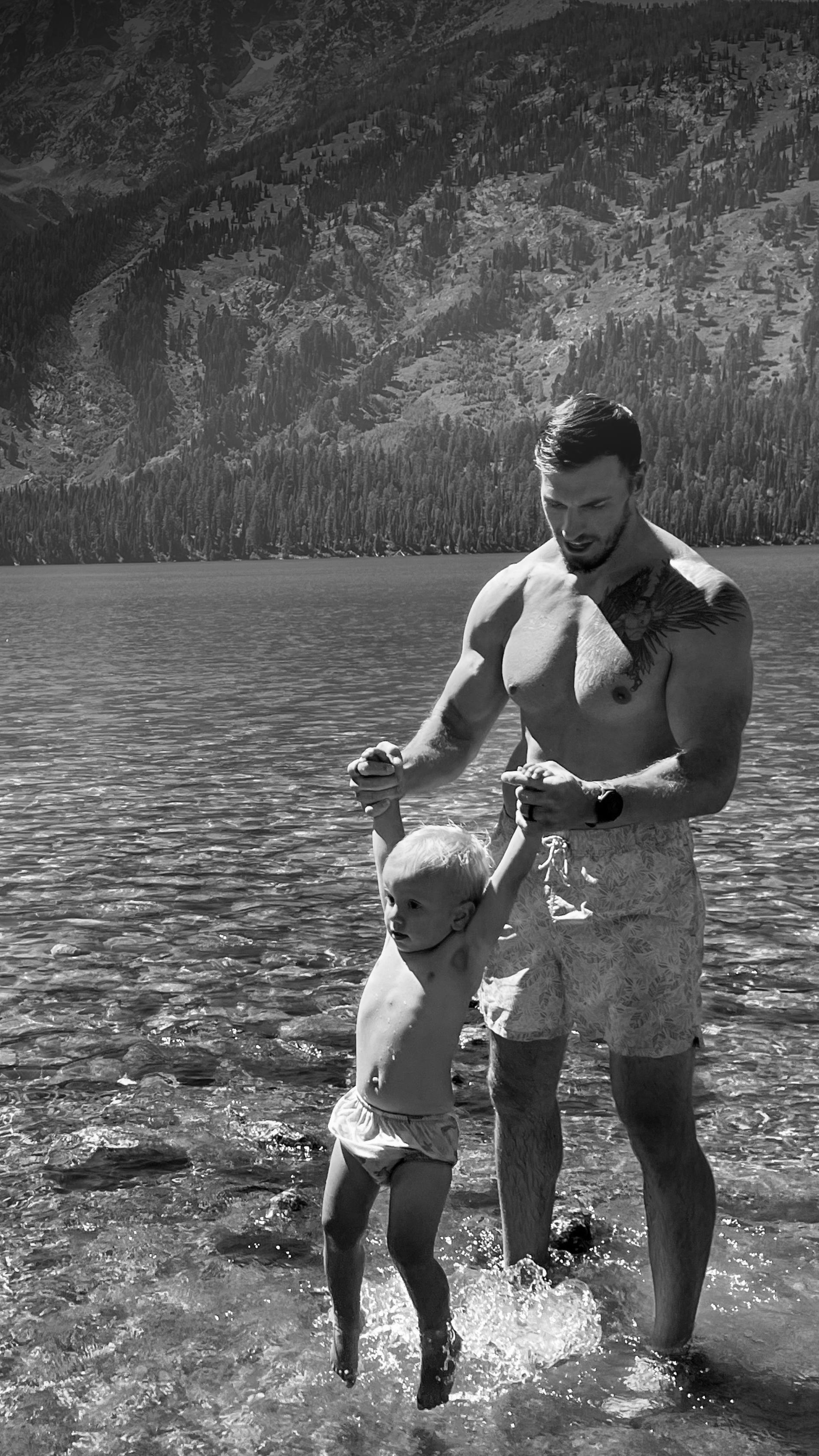 A man and a young child playing in a lake with mountains and forests in the background.