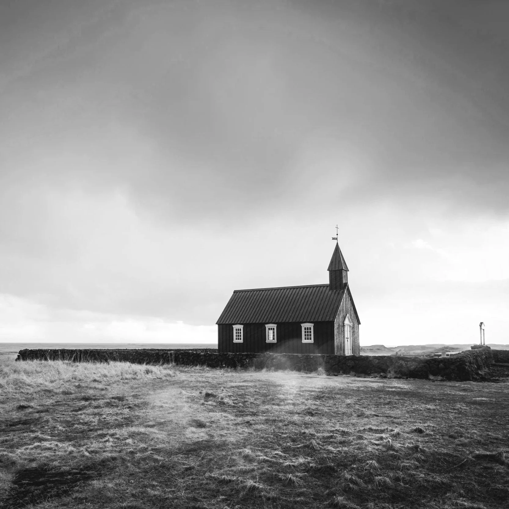 A small wooden church with a steeple on a rocky landscape, under a cloudy sky, in black and white.