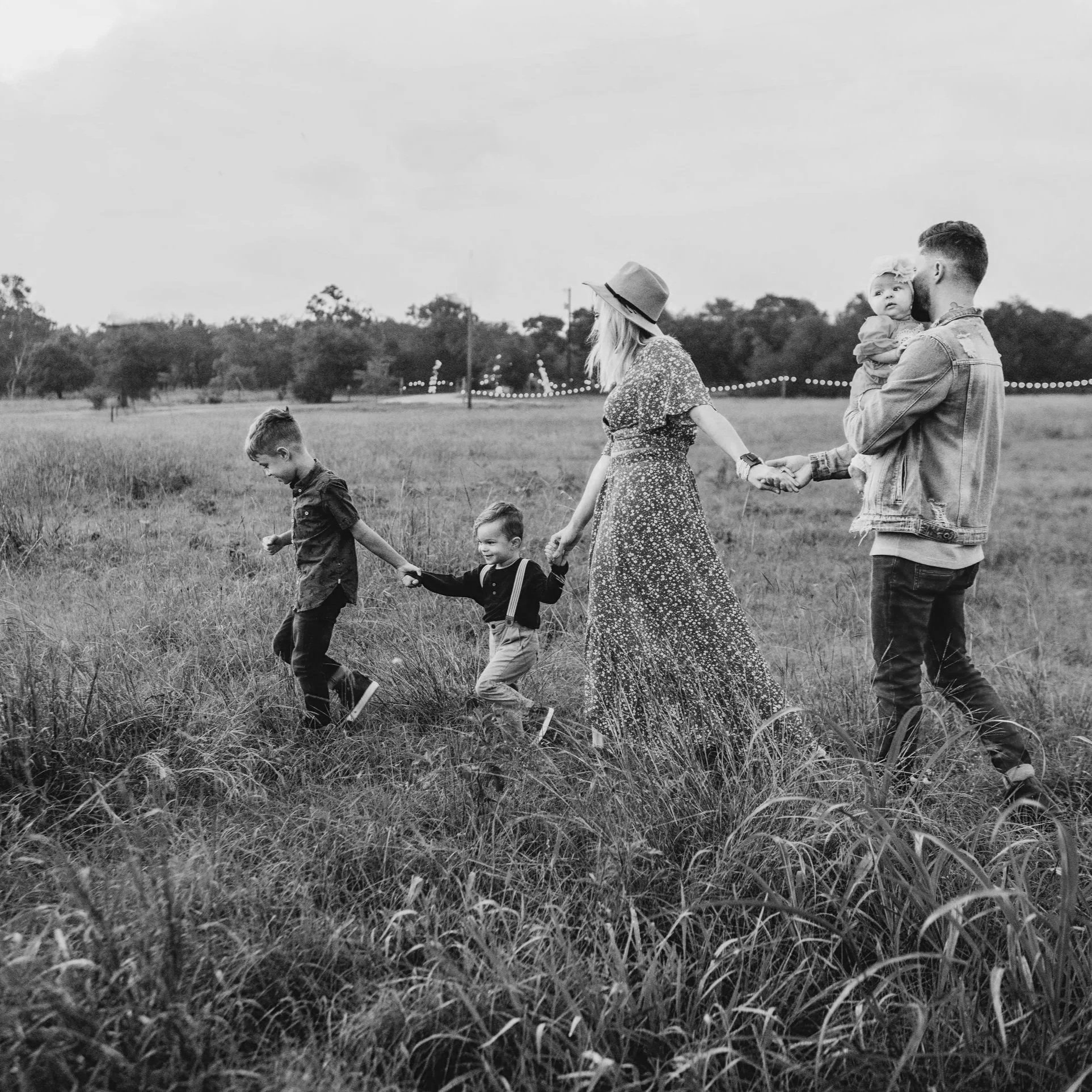 A black and white photo of a family walking through a grassy field. The group includes two men, two women, and three children. One man is holding a young girl, and the woman in the middle is holding the hand of a young boy. The children are walking ahead of the adults.