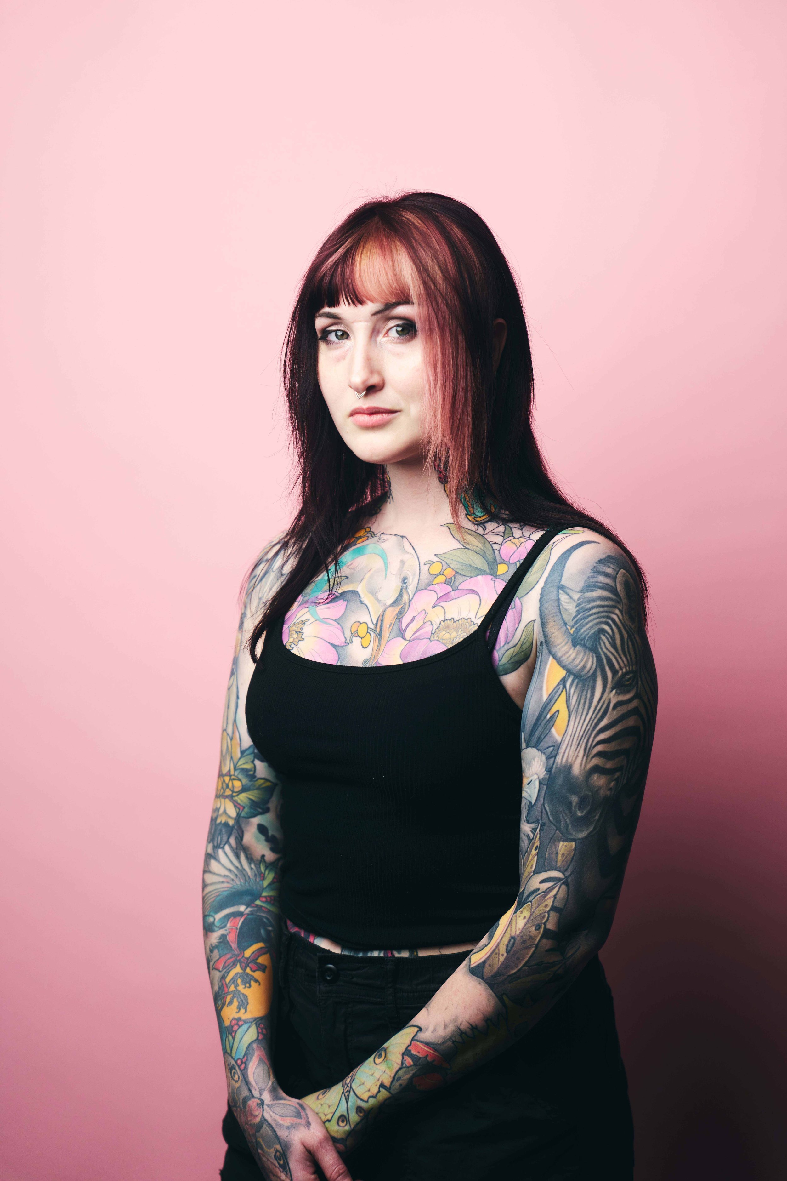 A woman with pink and black hair, tattoos on both arms and upper chest, wearing a black tank top, standing against a light pink background.