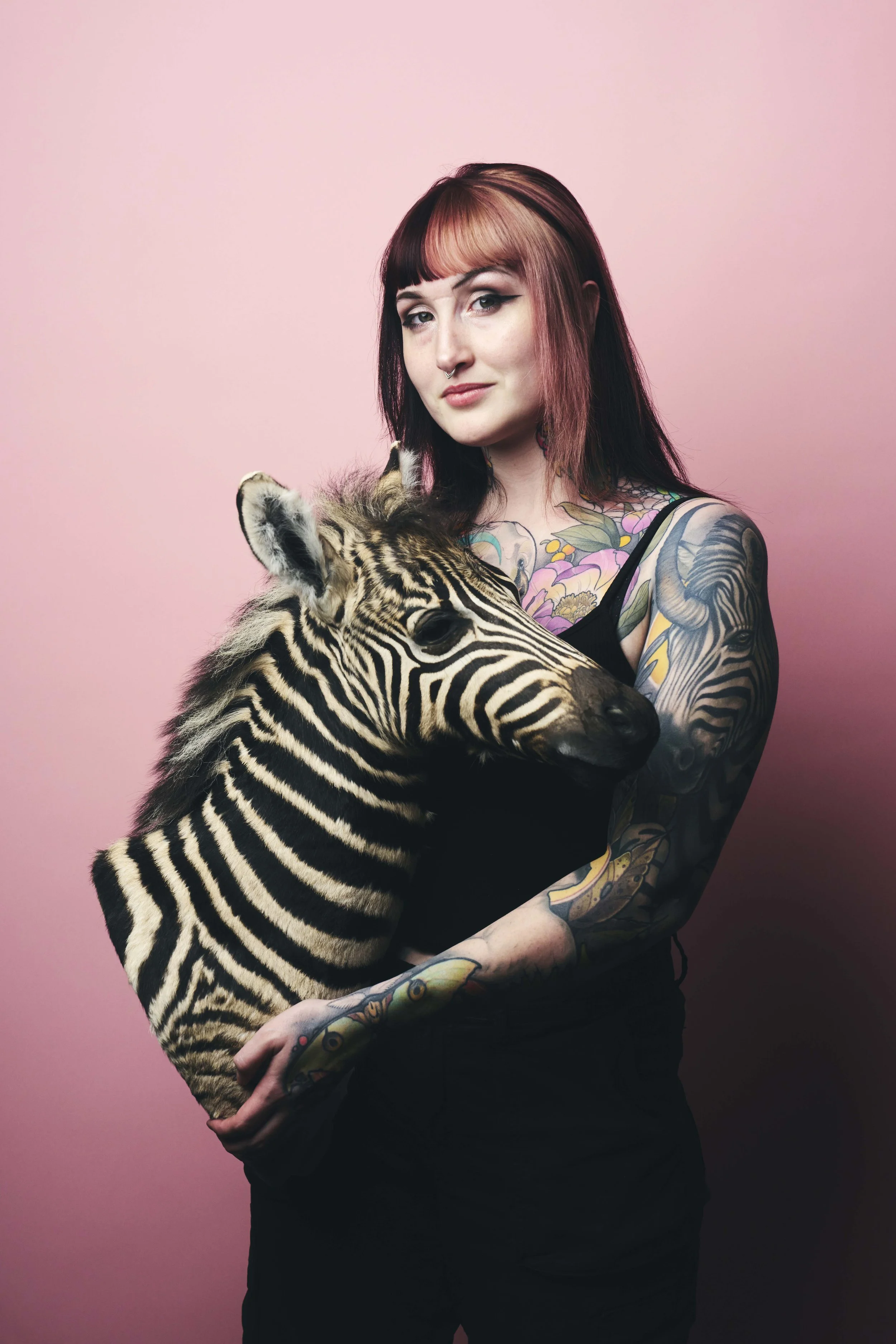 A woman with pink and black hair and tattoos holding a baby zebra against a pink background.