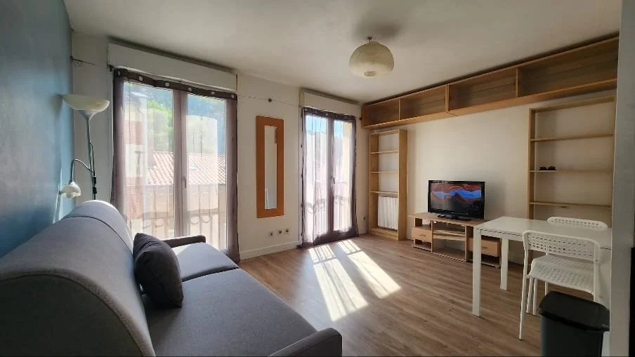 Salon avec canapé gris, télévision, étagères en bois, table blanche, lampadaire et deux portes-fenêtres avec rideaux laissant entrer la lumière