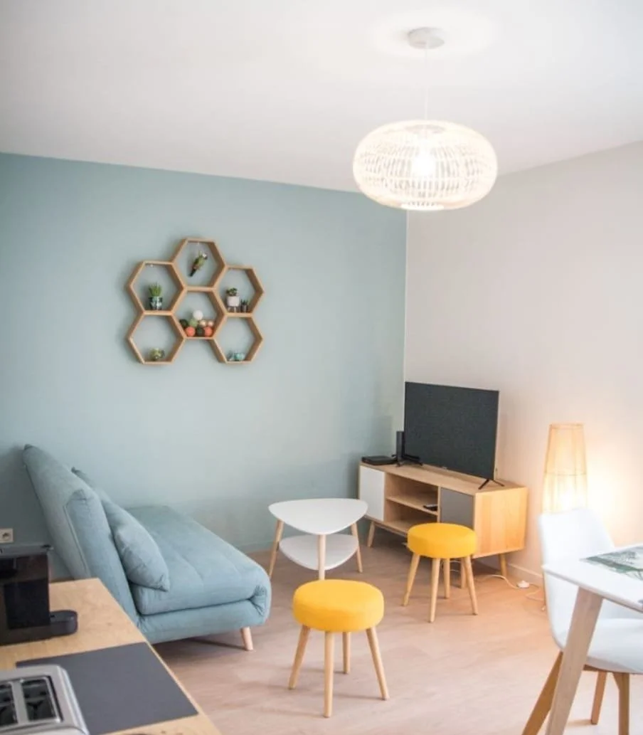 Salon moderne avec canapé bleu, télévision sur meuble en bois, lampe sur le côté, table blanche, poufs jaunes, et étagères en forme de hexagone sur le mur.