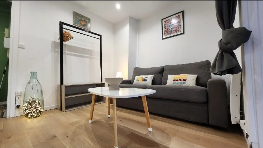 Salon moderne avec canapé gris, table basse ronde blanche, décorations sur le mur, lampadaire et grande jarre avec lumière, fenêtre avec rideau noir.