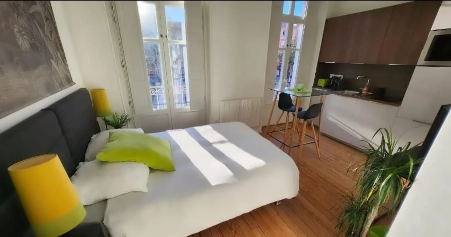 Chambre d'un studio avec lit, deux lampes jaunes, une plante verte, fenêtre laissant entrer la lumière, coin cuisine avec table et chaises.