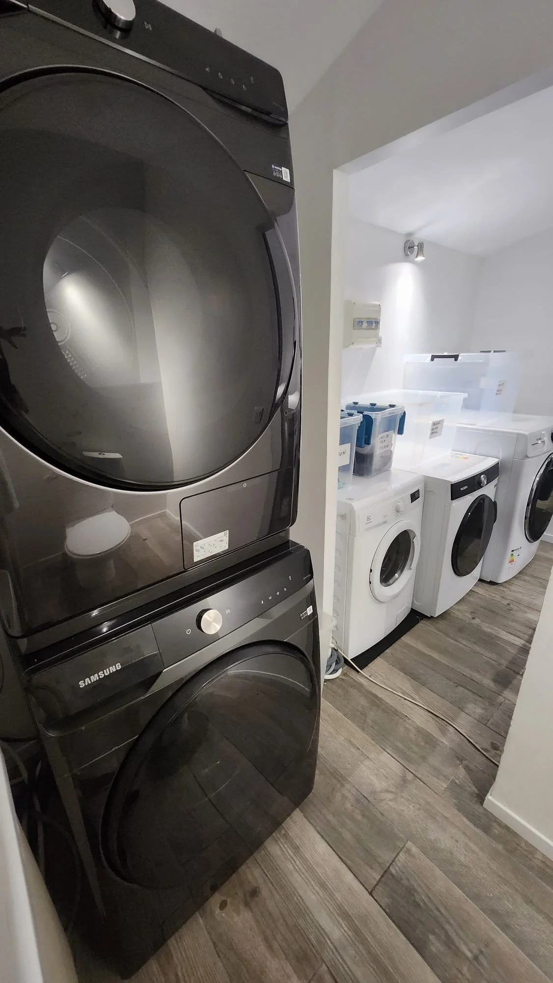 Une buanderie avec une machine à laver et un sèche-linge noirs empilés, ainsi que plusieurs autres appareils de lavage blancs alignés dans une pièce bien éclairée.