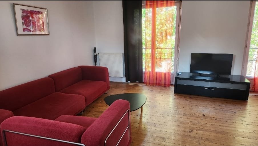 Salon avec canapé rouge, télévision noire sur meuble, fenêtres avec rideaux orange, parquet en bois, tableau sur le mur, radiateur et porte voilée.