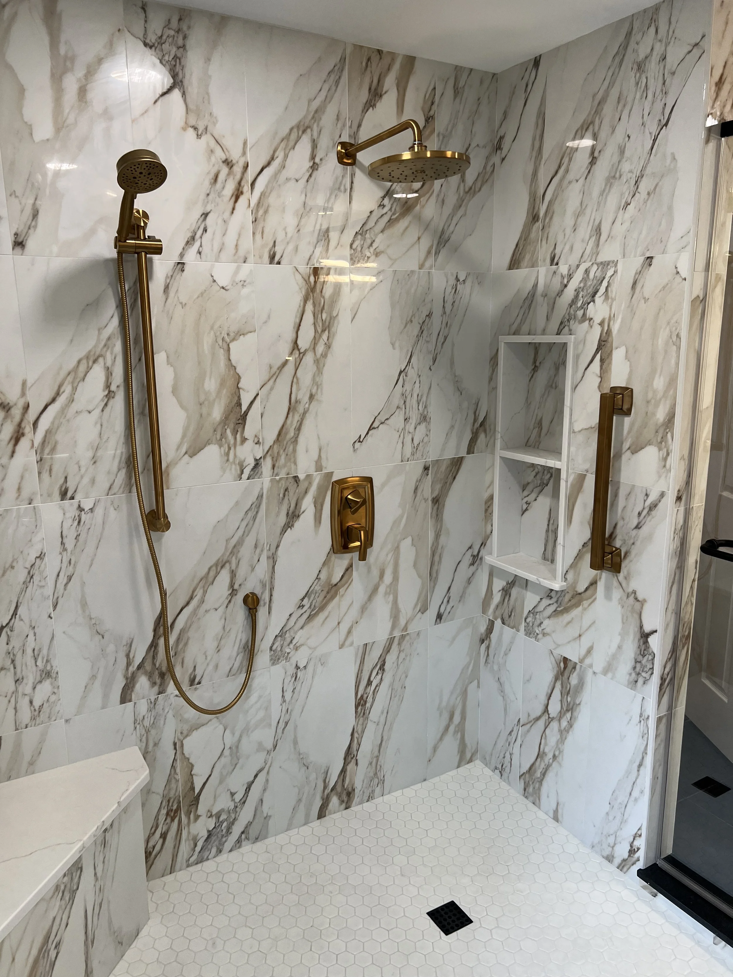 Modern walk-in shower with marble wall tiles, gold fixtures, a handheld showerhead, and a ceiling-mounted rainfall showerhead. The shower has a built-in niche and a white hexagonal tile floor.