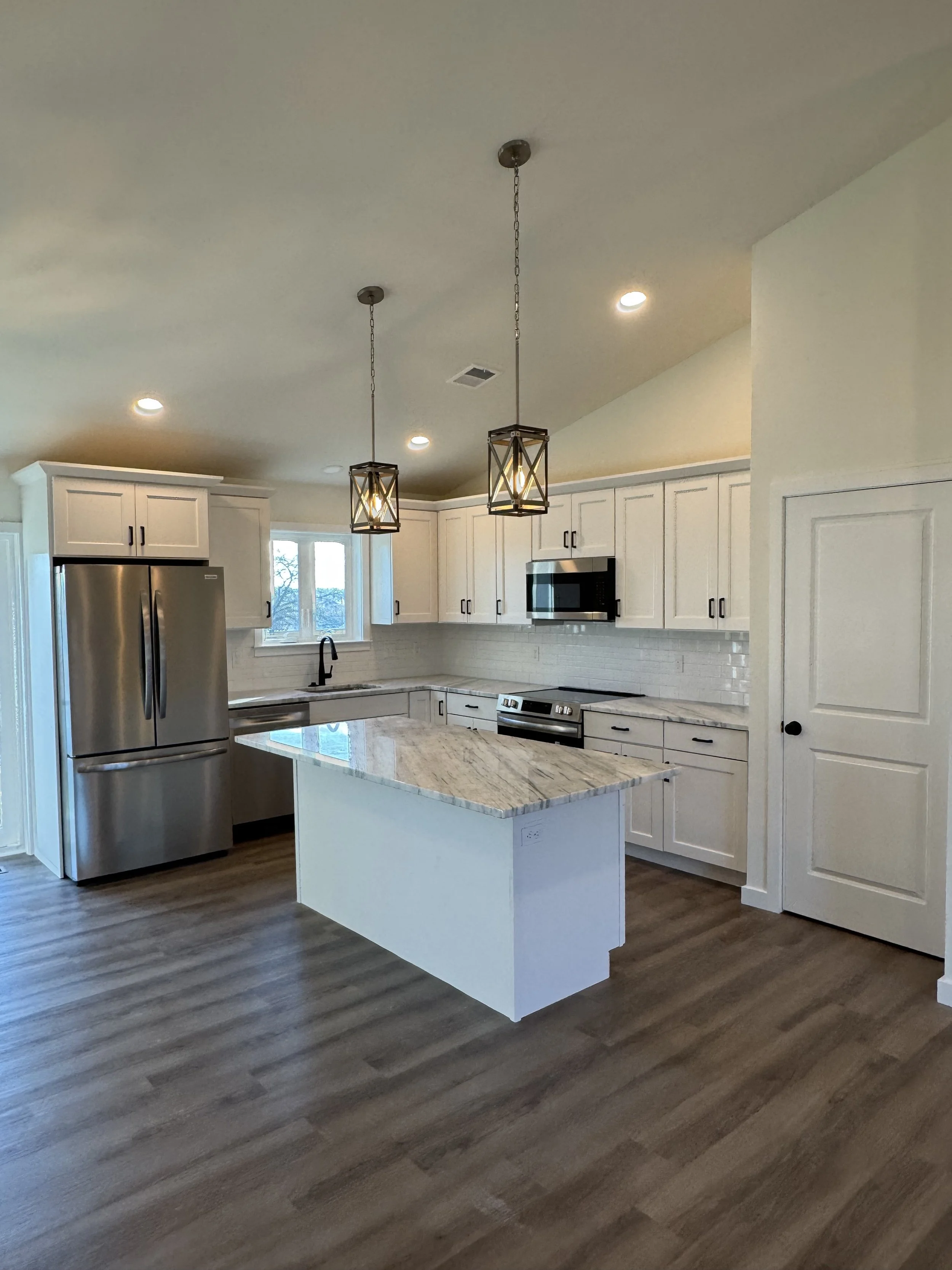 Modern kitchen with white cabinets, stainless steel appliances, a marble island, and two pendant lights hanging from a high ceiling. Hardwood floors and a window above the sink are visible.