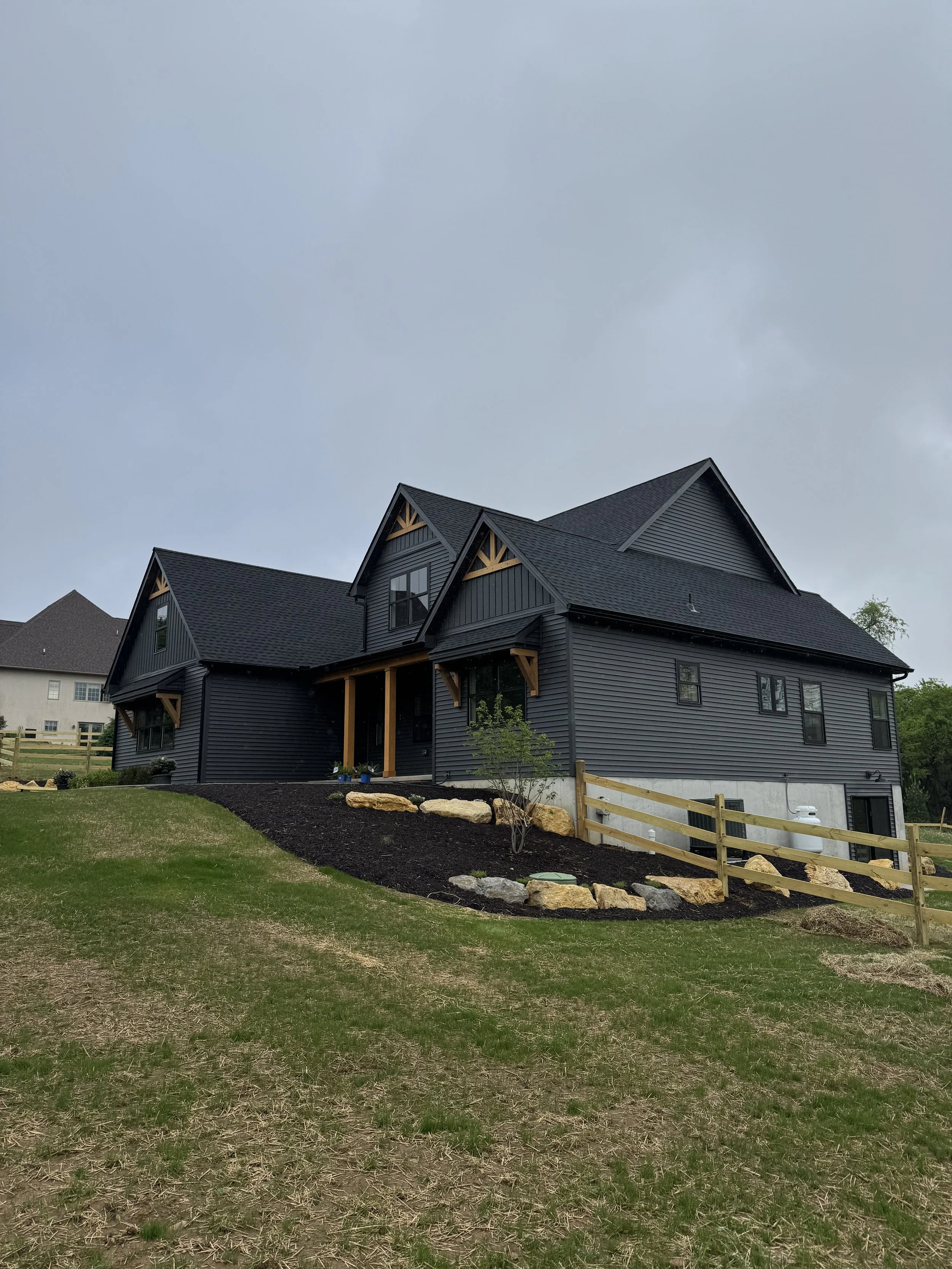 A dark blue house with a steep gabled roof, multiple windows, and a small porch, surrounded by a grassy yard with some rocks and a wooden fence, under a cloudy sky.