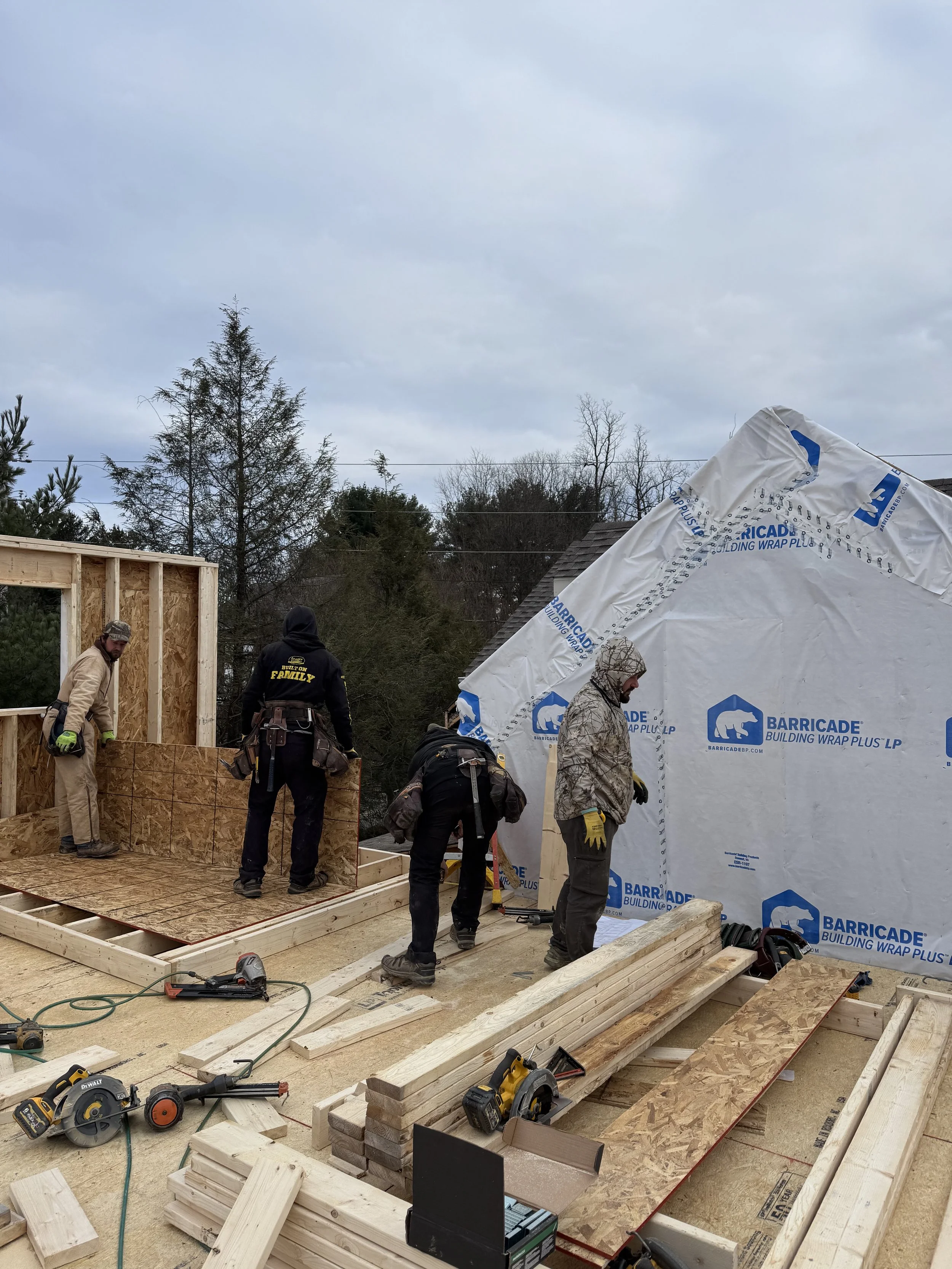 Construction workers building a wooden structure, with framing and tools on site, and a partially covered wall using building wrap.