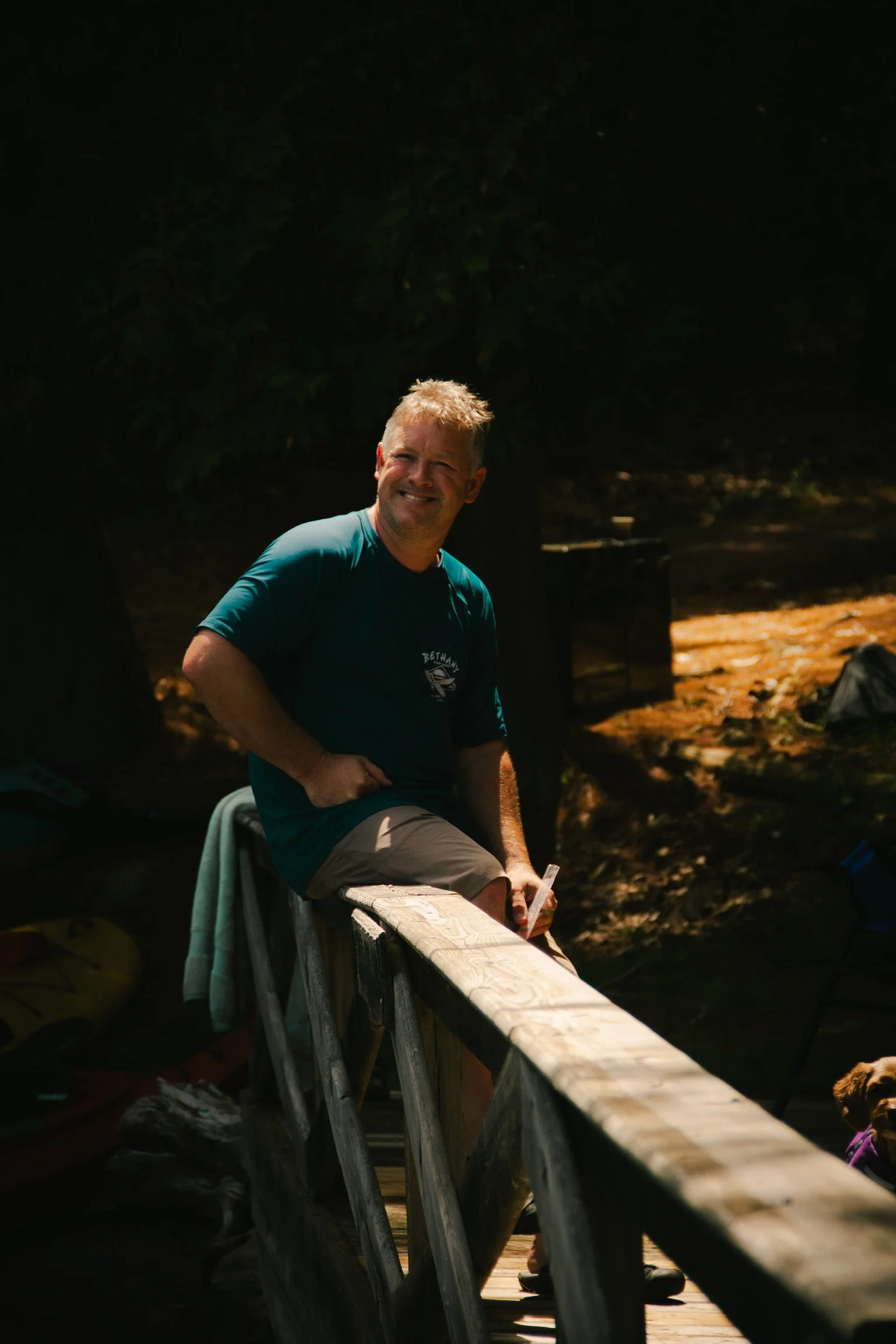 A man sitting on a wooden railing outdoors, smiling,, with trees in the background and shadows cast by sunlight.