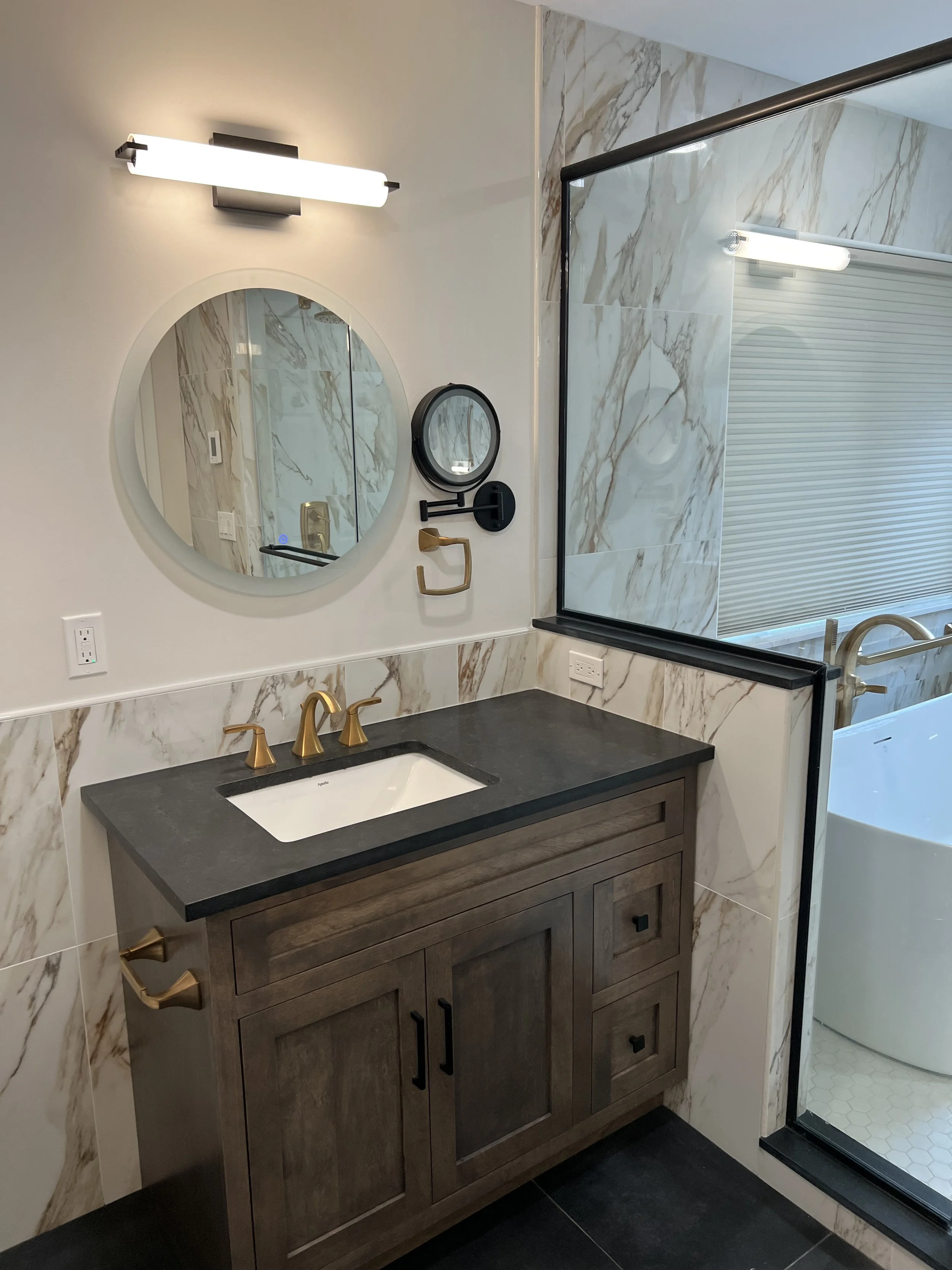 Bathroom vanity with a wooden cabinet, black countertop, gold faucet, oval mirror, black wall mirror, and glass shower enclosure with marble tiles.