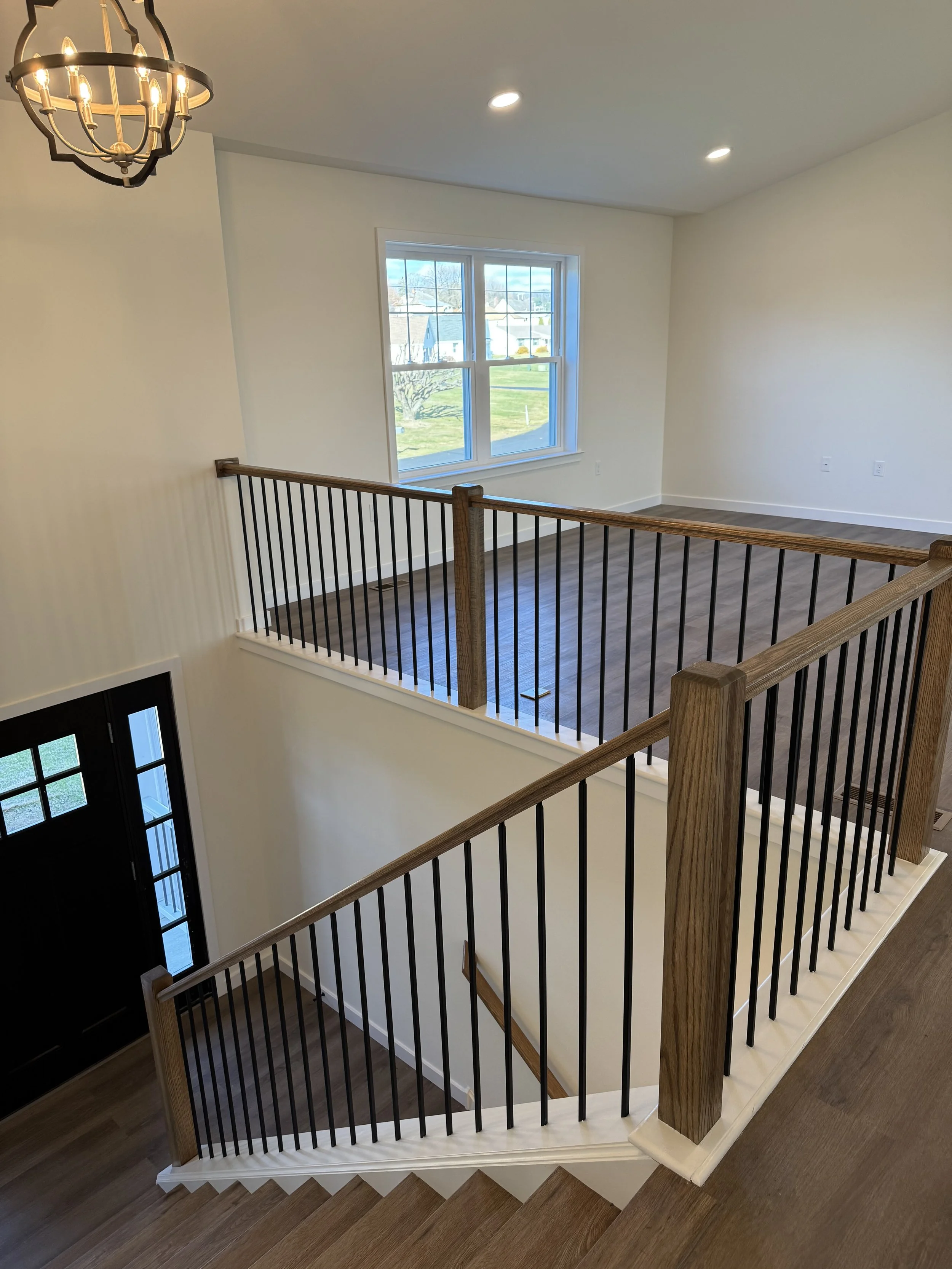 View of a staircase landing in a house, with a wooden handrail and black metal balusters, a large window showing the outdoor yard, and a black front door with glass panels. The ceiling has recessed lighting and a hanging chandelier.