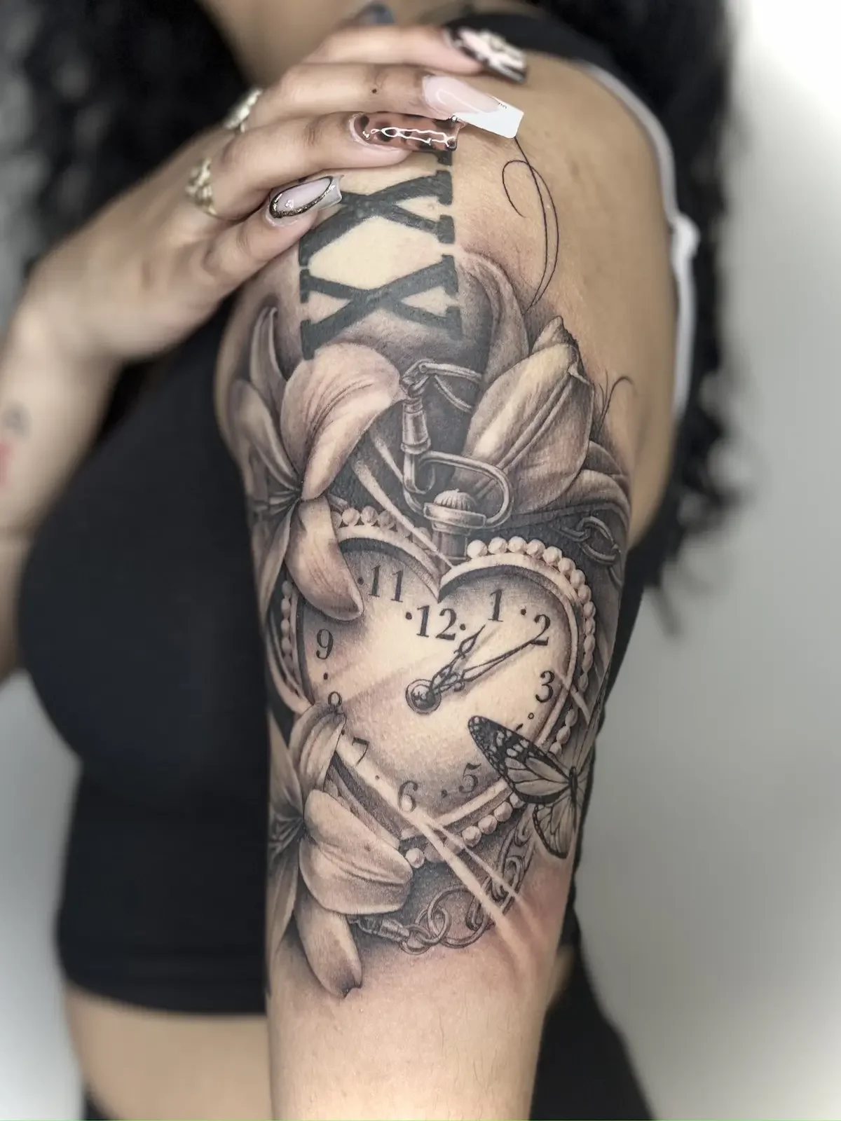 Black and grey realism upper arm tattoo with heart clock, lilies, and butterfly by Johny Vasquez — Mommasboy Tattoo Austin