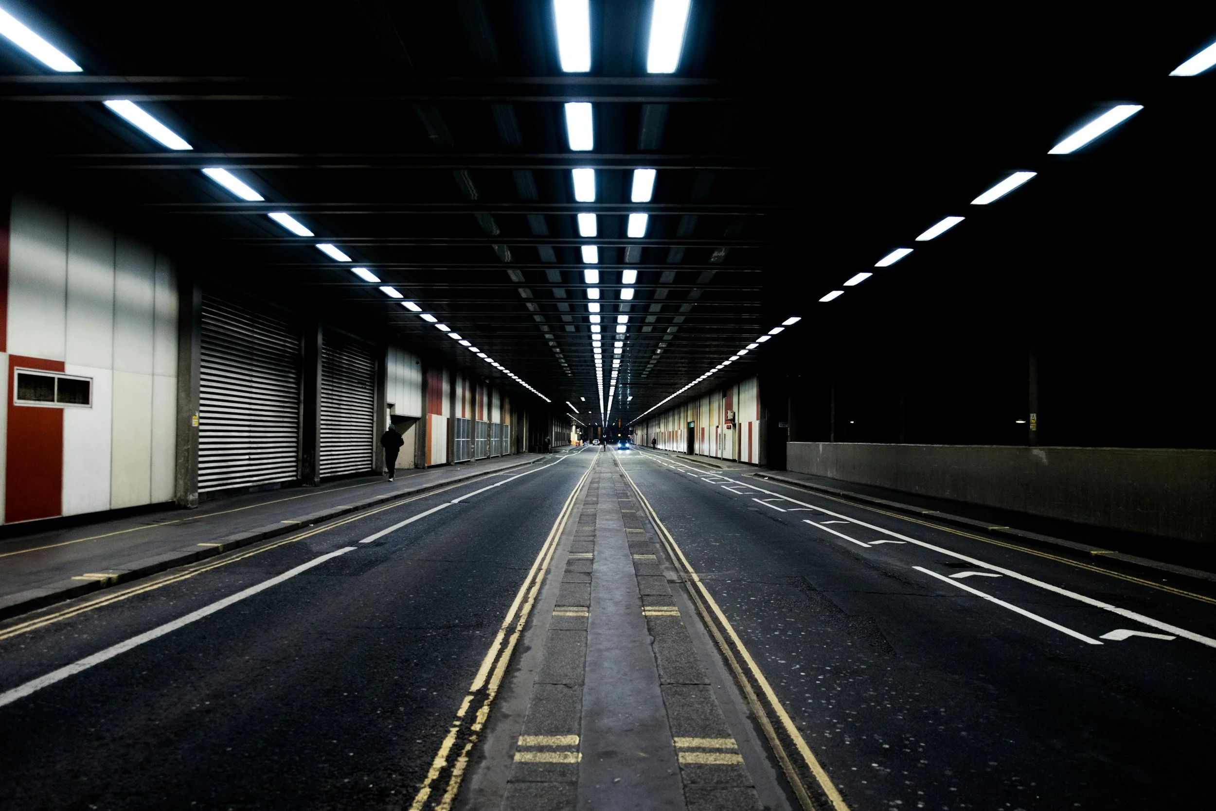 Empty city street under a bridge or tunnel, with road markings and a person walking on the left sidewalk at night.