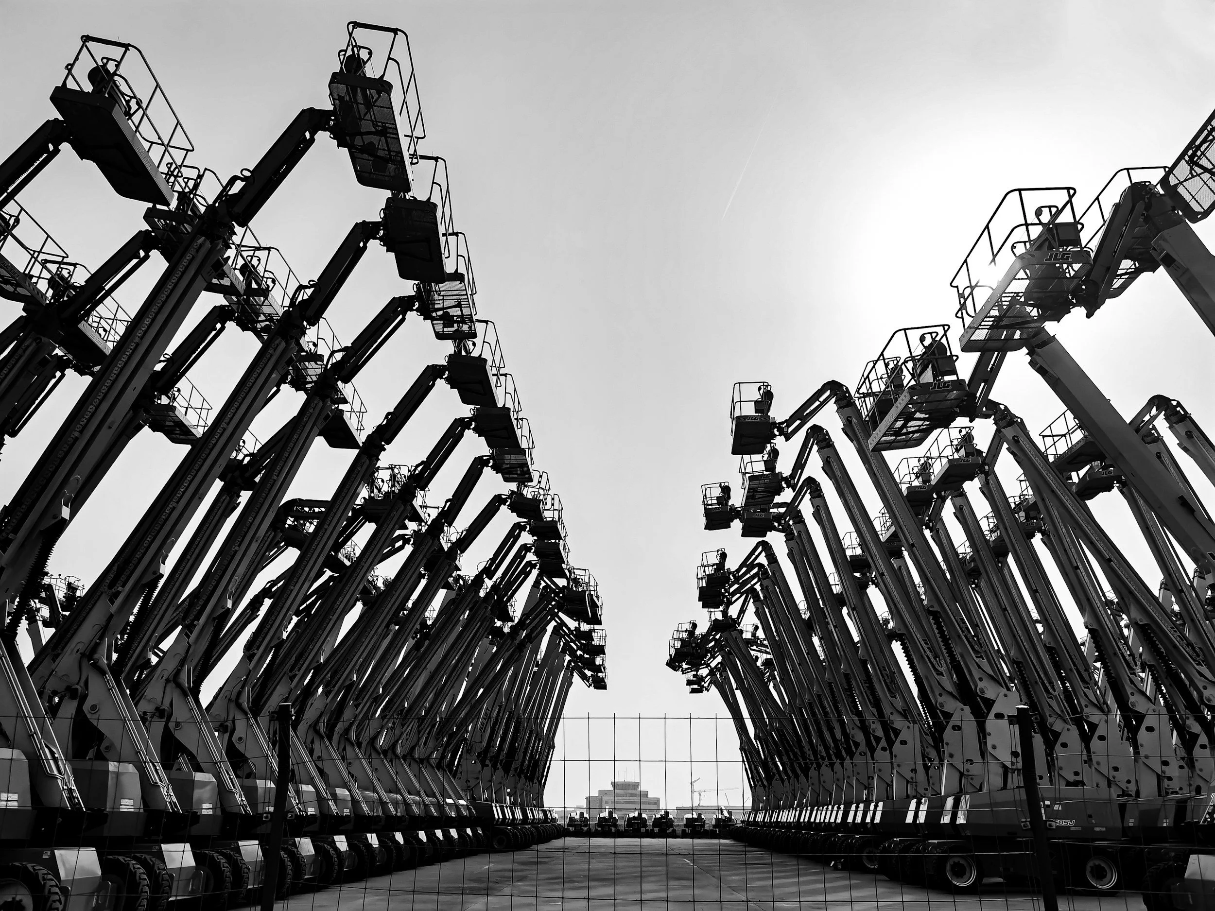 A black and white photo of multiple crane lifts lined up in two rows at a construction yard with a chain-link fence in the foreground.