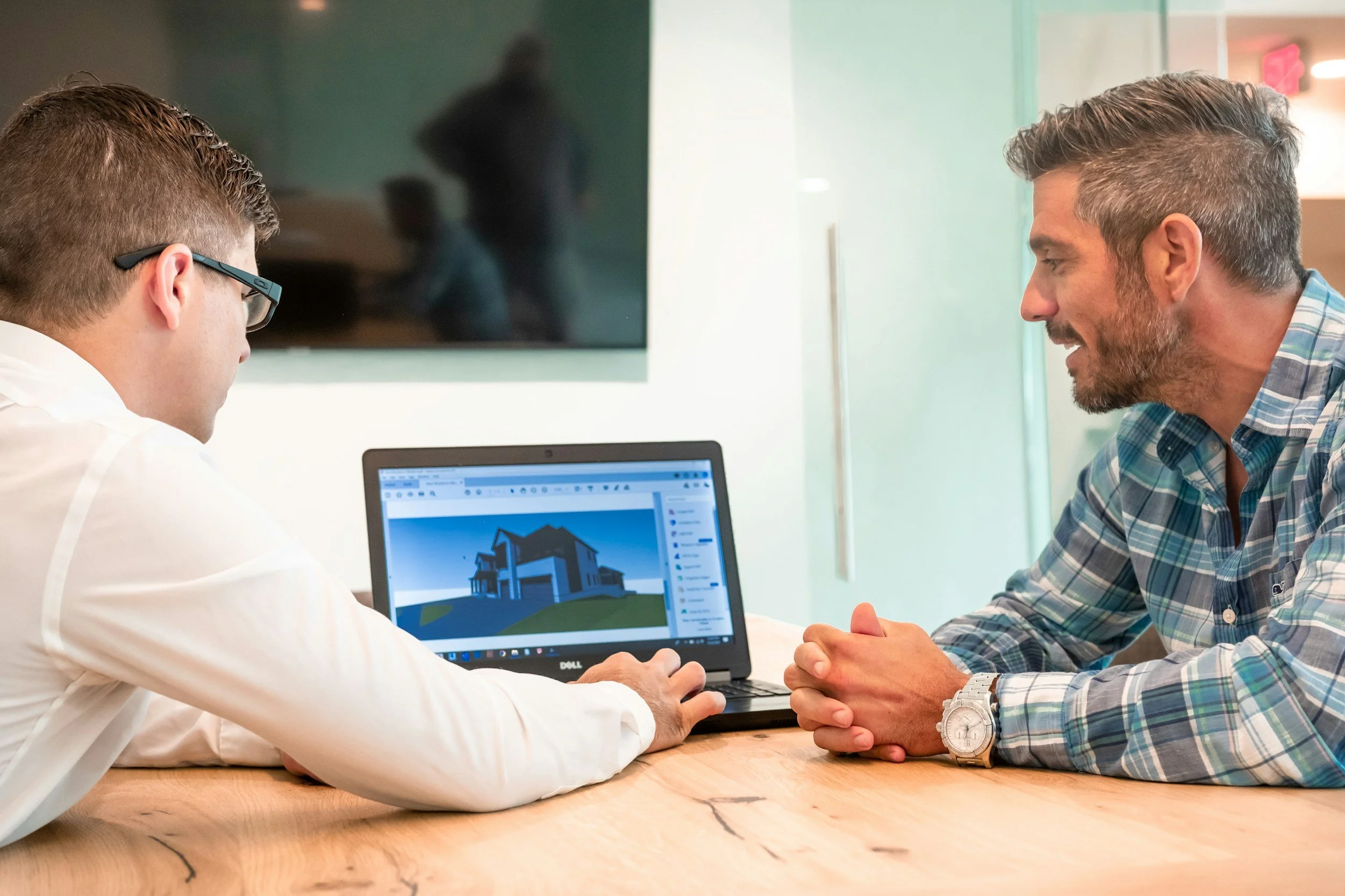 Two men sitting at a wooden table, discussing house design on a laptop, with a large TV screen in the background.