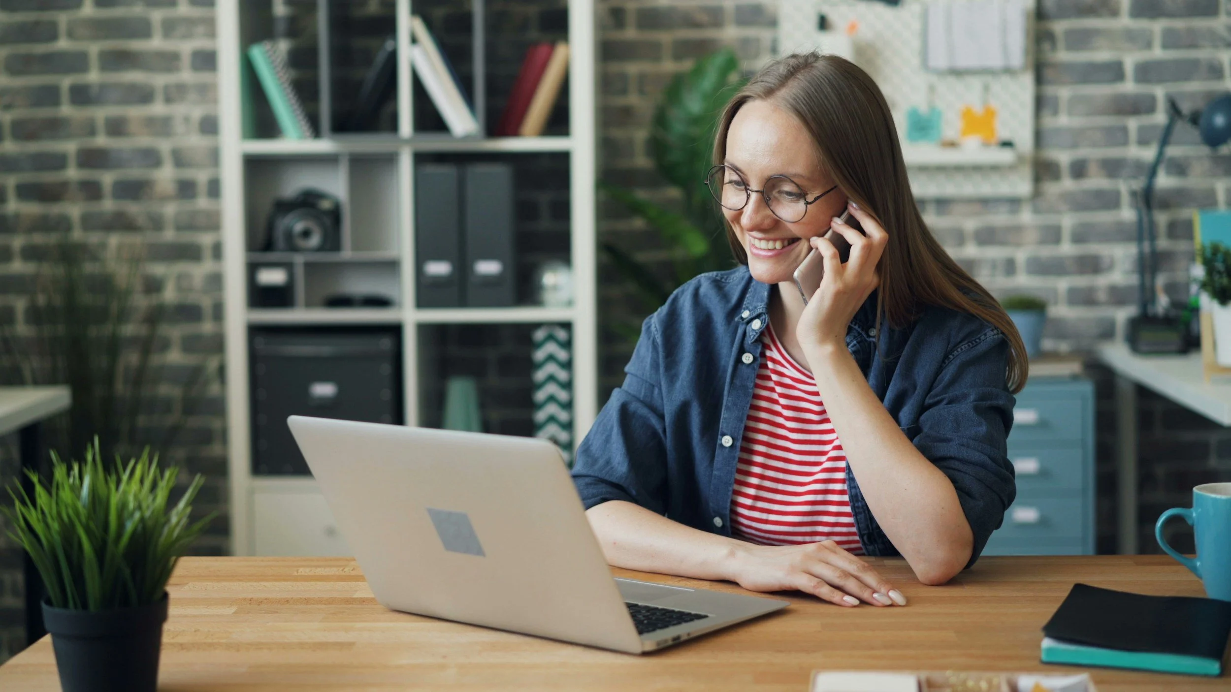 A woman with long brown hair and glasses smiling while talking on her cellphone at her desk with a laptop and a blue mug, in a modern office with a brick wall and a bookshelf in the background.