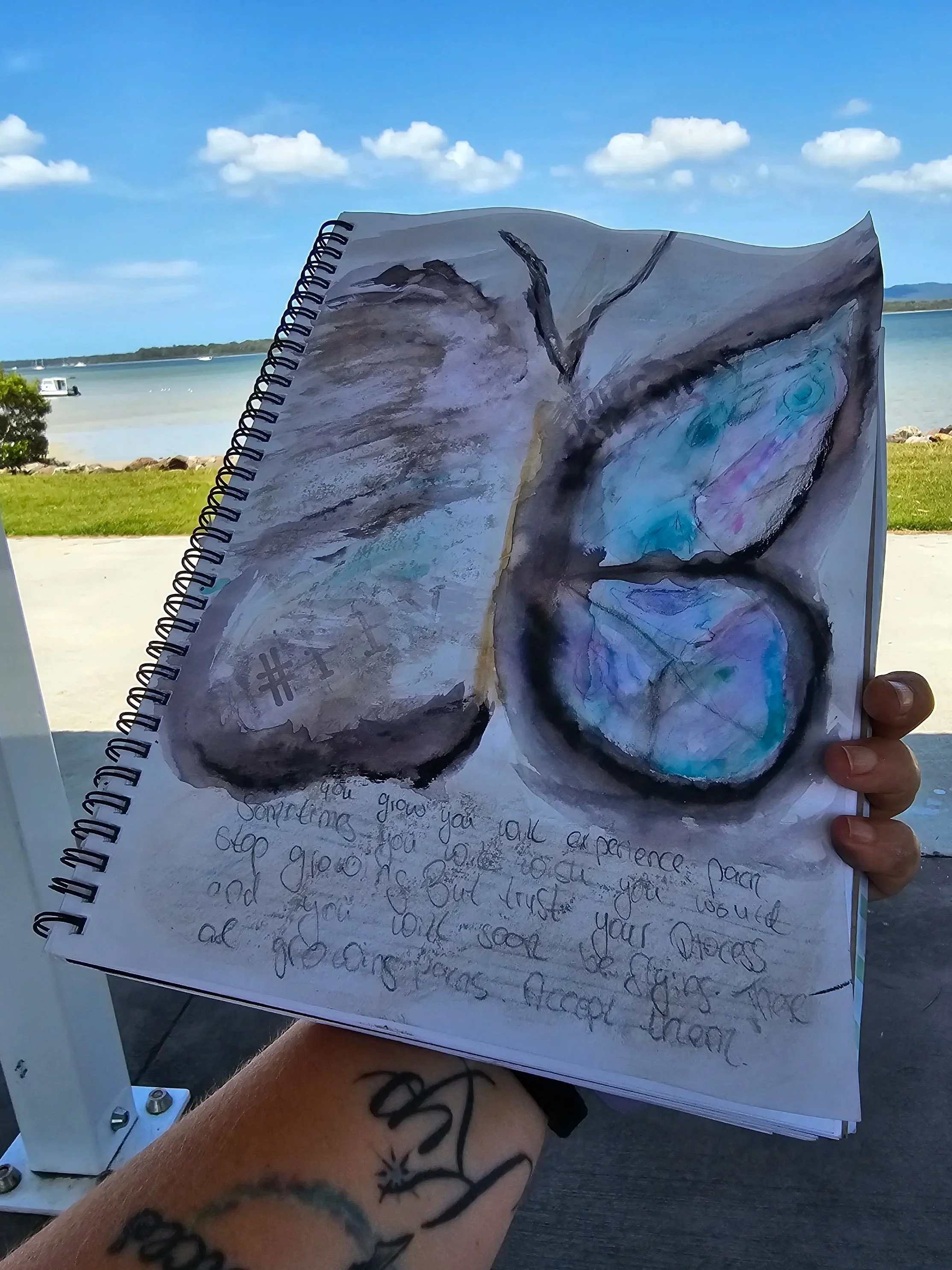 A hand holding a spiral notebook with a watercolor painting of a pair of glasses and an eye. The background shows a body of water, boats, blue sky, and some clouds.