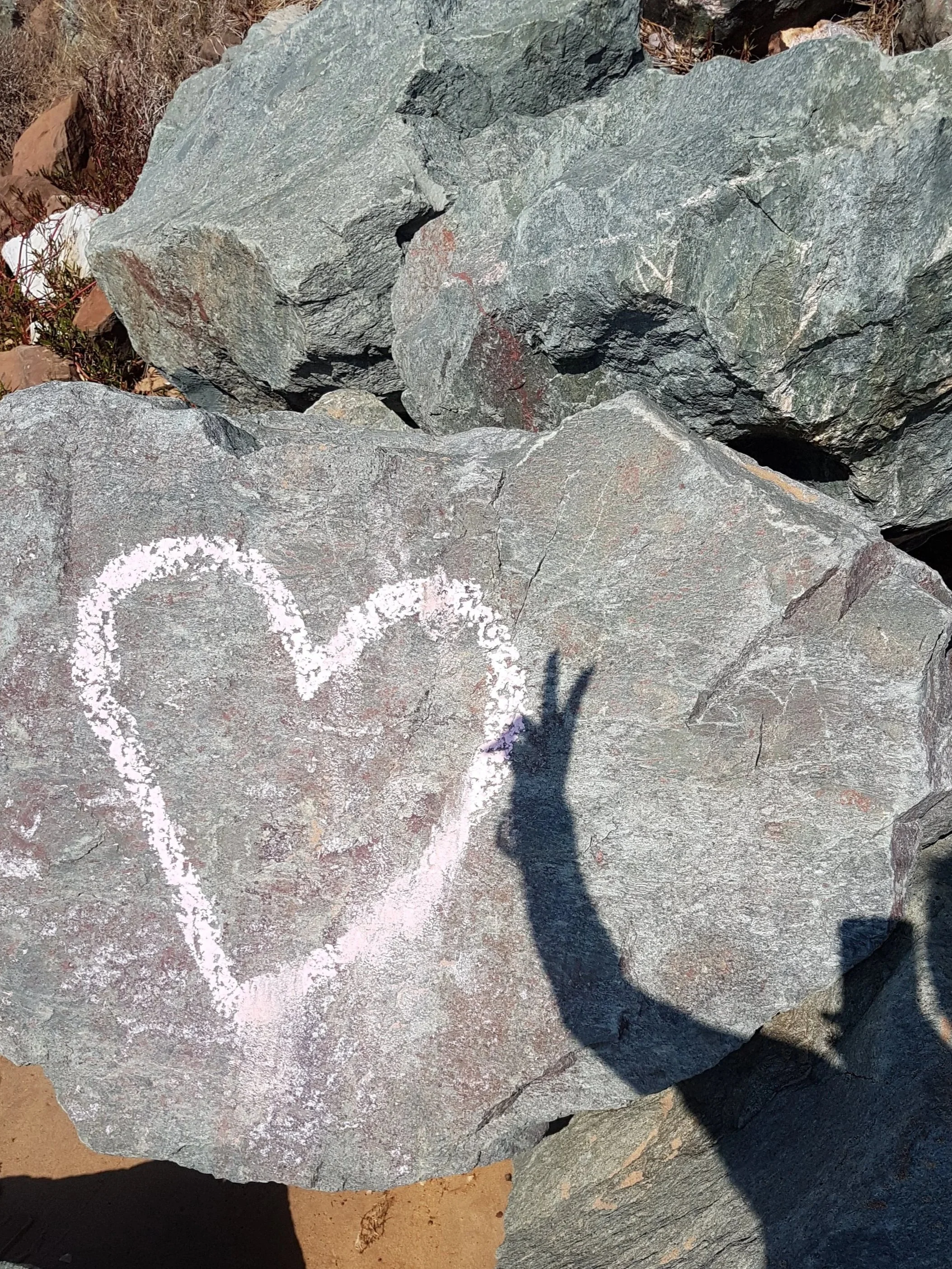 Large gray rocks on the ground with a chalk painted love heart and a persons shadow with the peace sign