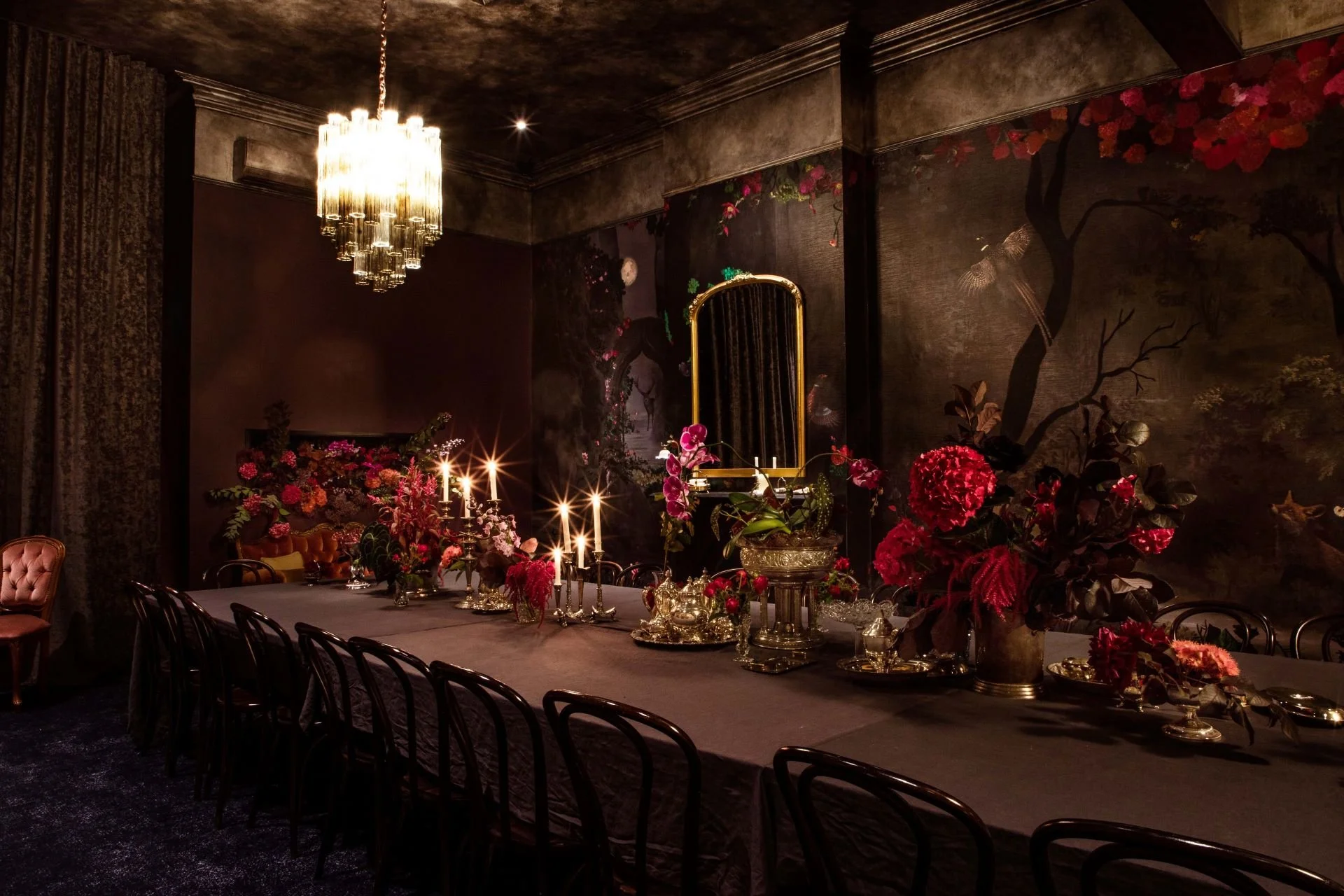 Elegant private dining with a long table decorated with flowers and candles, ornate mirror, chandelier, and dark wall mural.