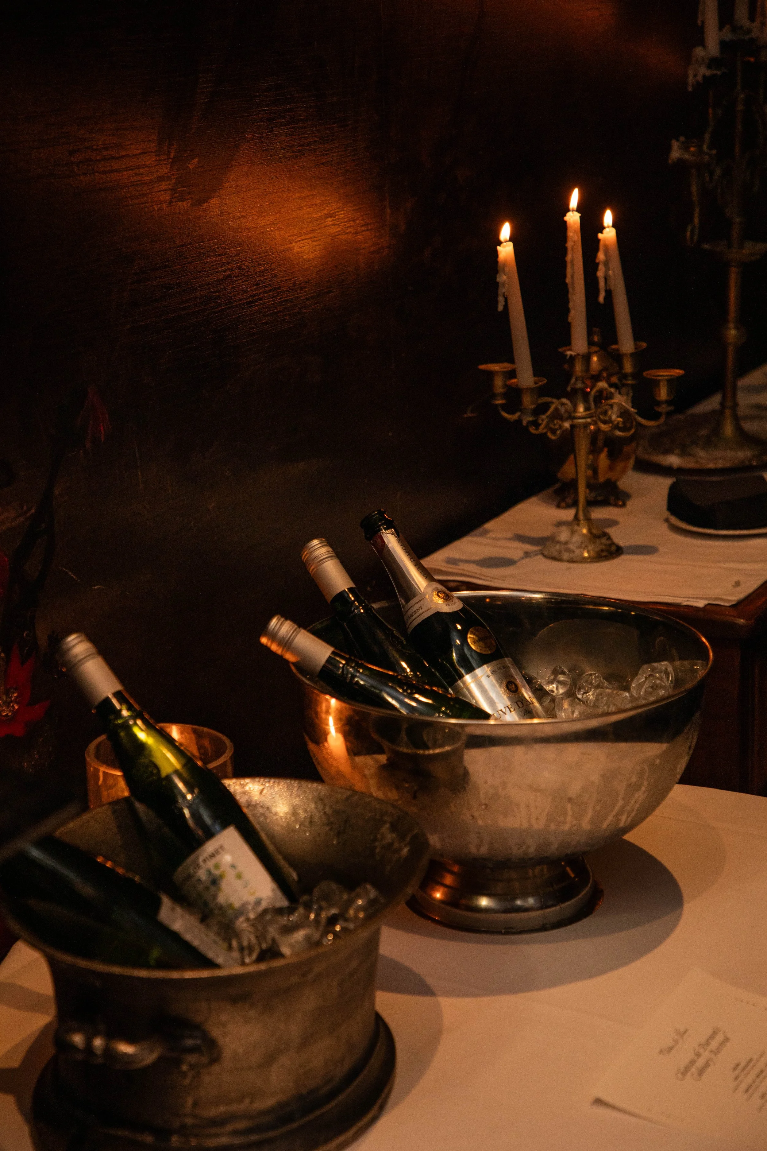 Bottles of wine in ice buckets, with a lit candelabra on a table in a dimly lit setting.