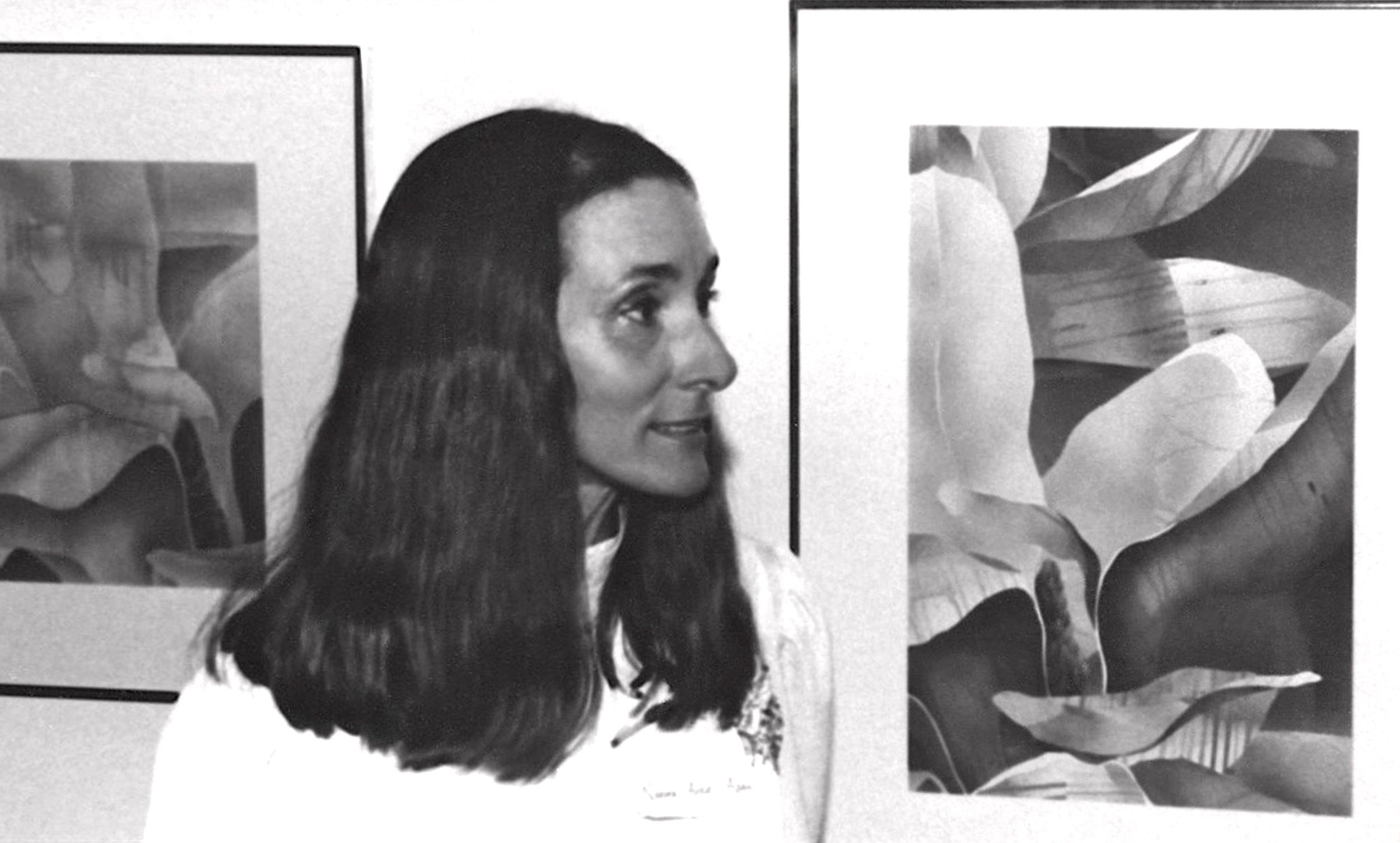 Black and white photograph of a woman with long dark hair profile view, standing in front of artwork including a painting of a face and large leaves.