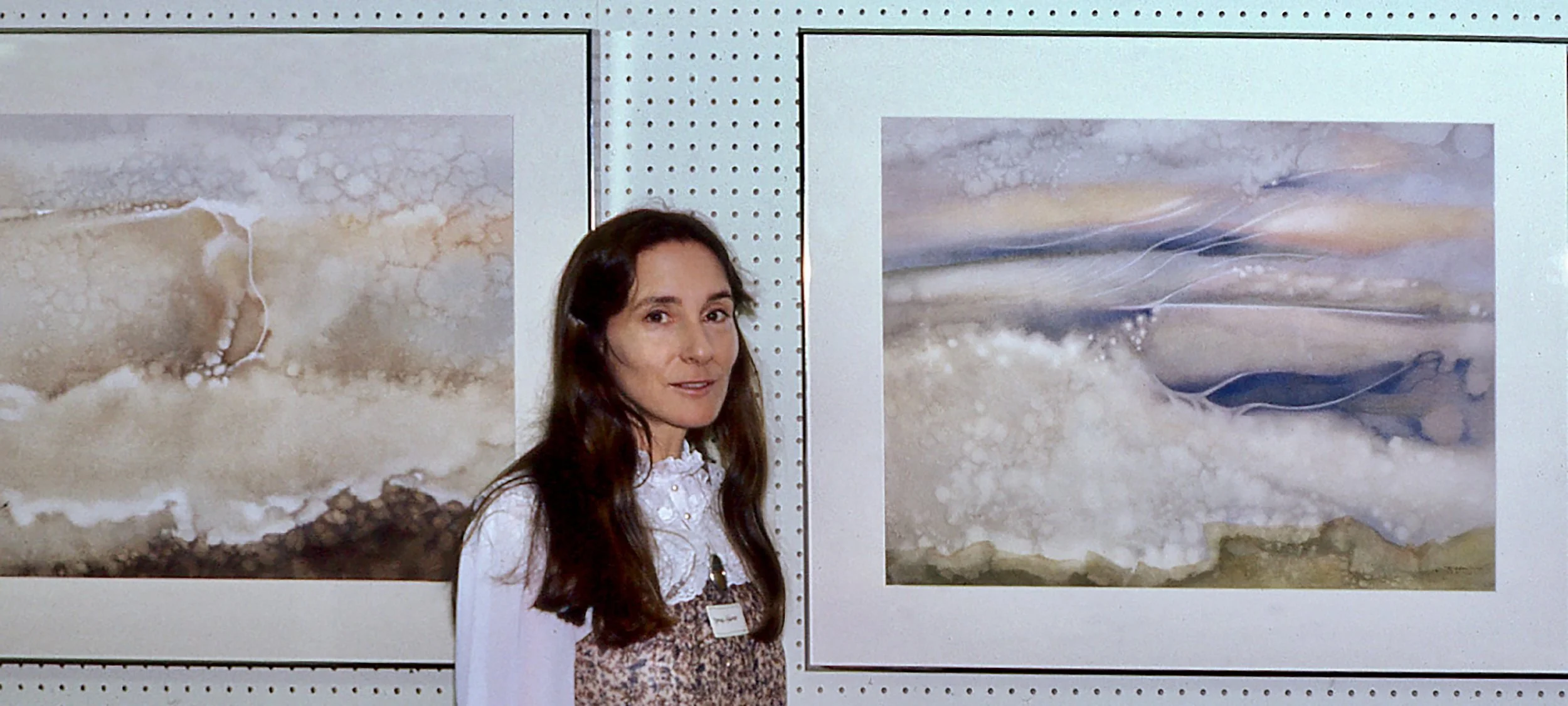 A woman with long dark hair and a patterned dress standing in front of abstract watercolor paintings at an art exhibition.