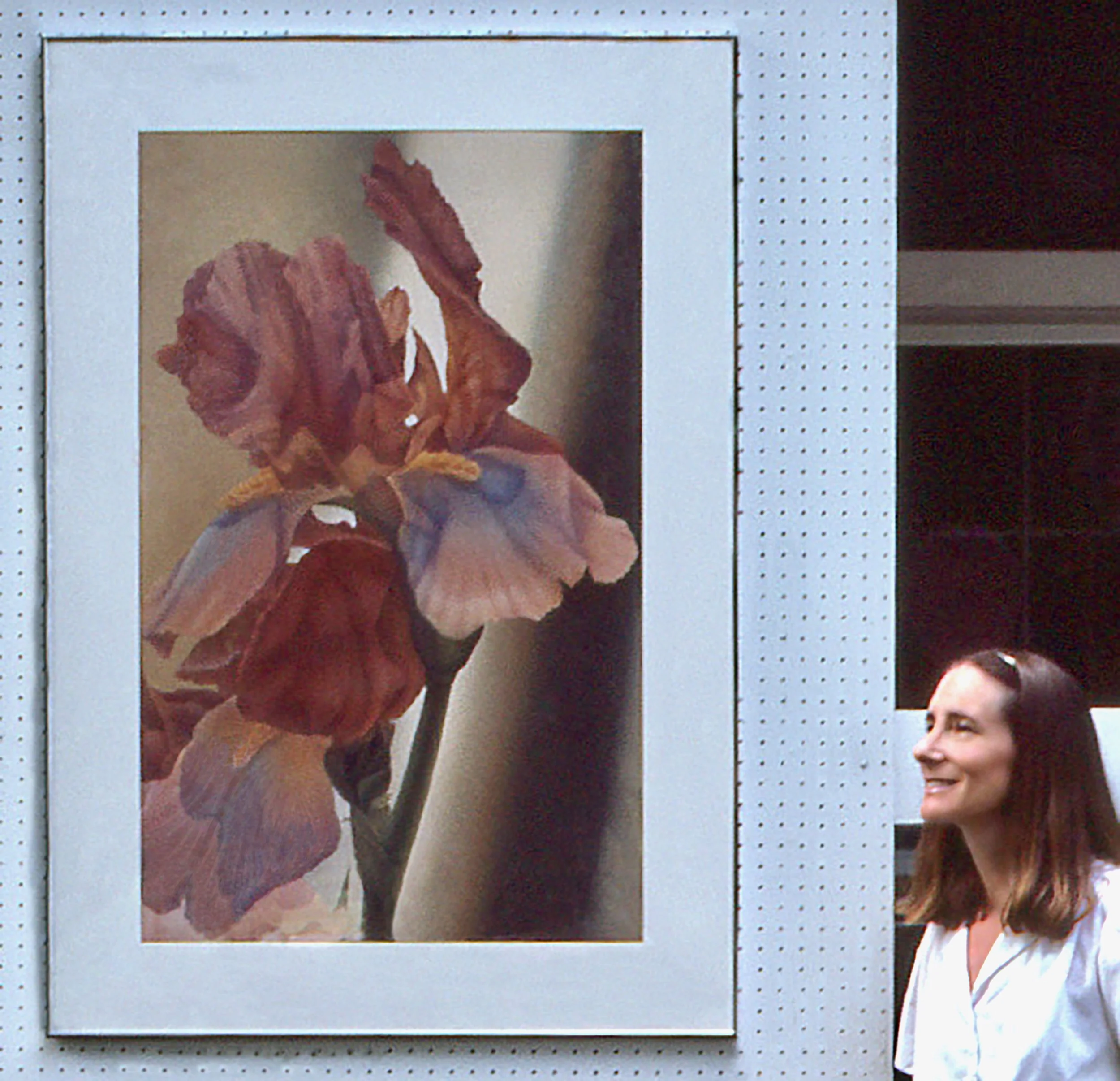 A woman smiling while viewing a framed painting of a colorful, detailed flower on a white wall.
