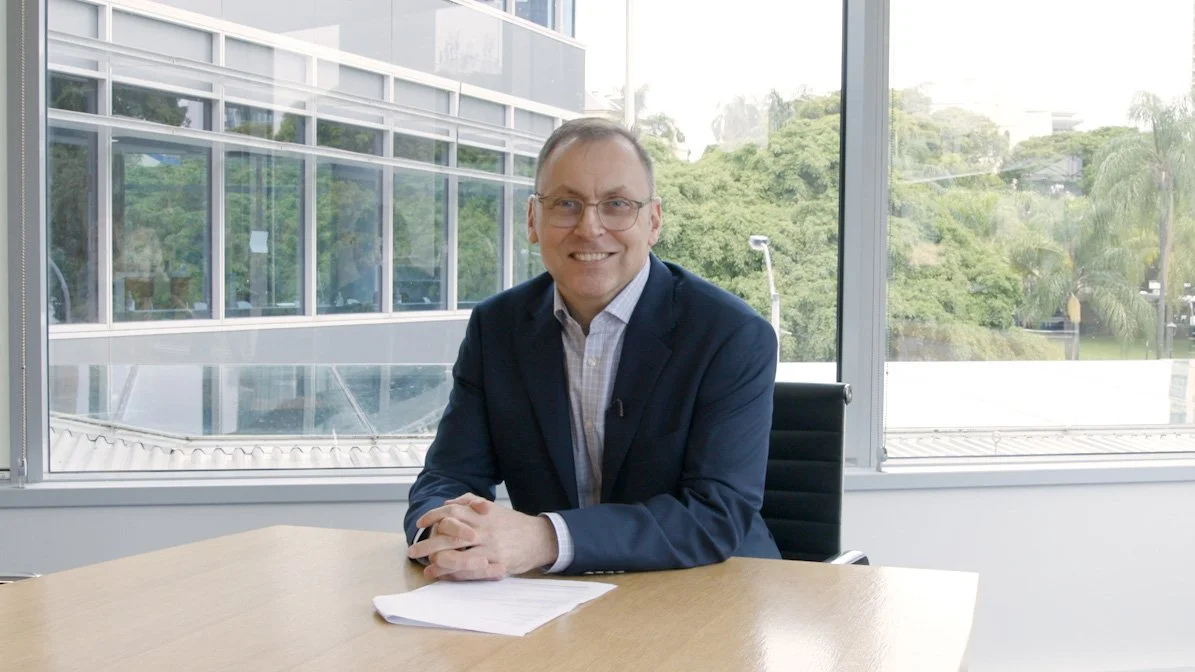 A smiling man in a suit sitting at a conference table in a modern office with large windows showing a green outdoor landscape.