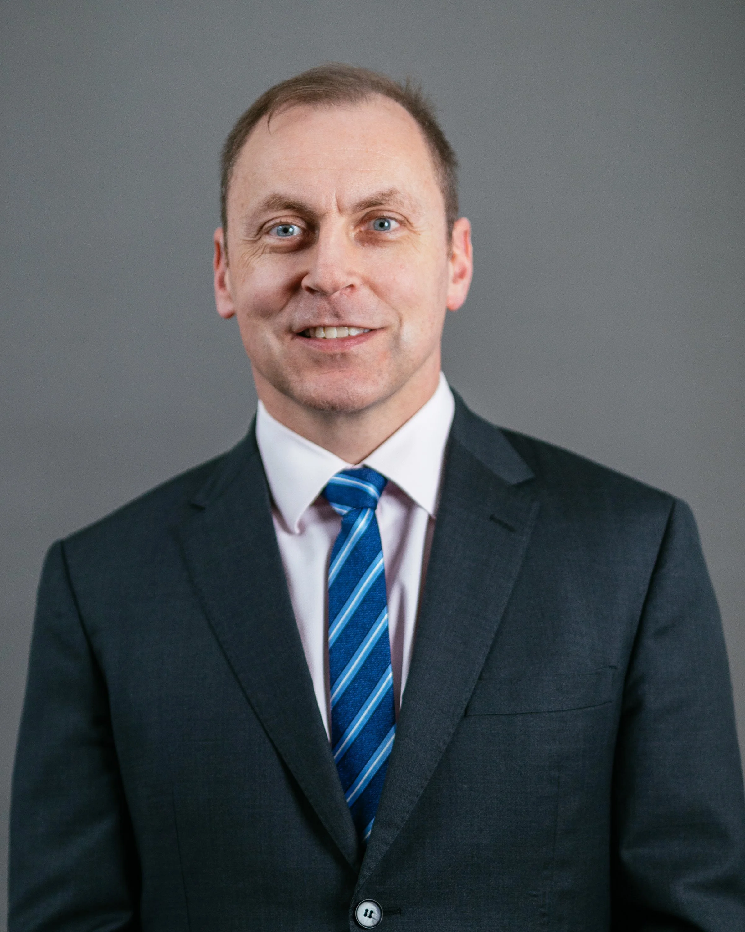 A man dressed in a dark suit, white shirt, and blue striped tie, smiling against a gray background.