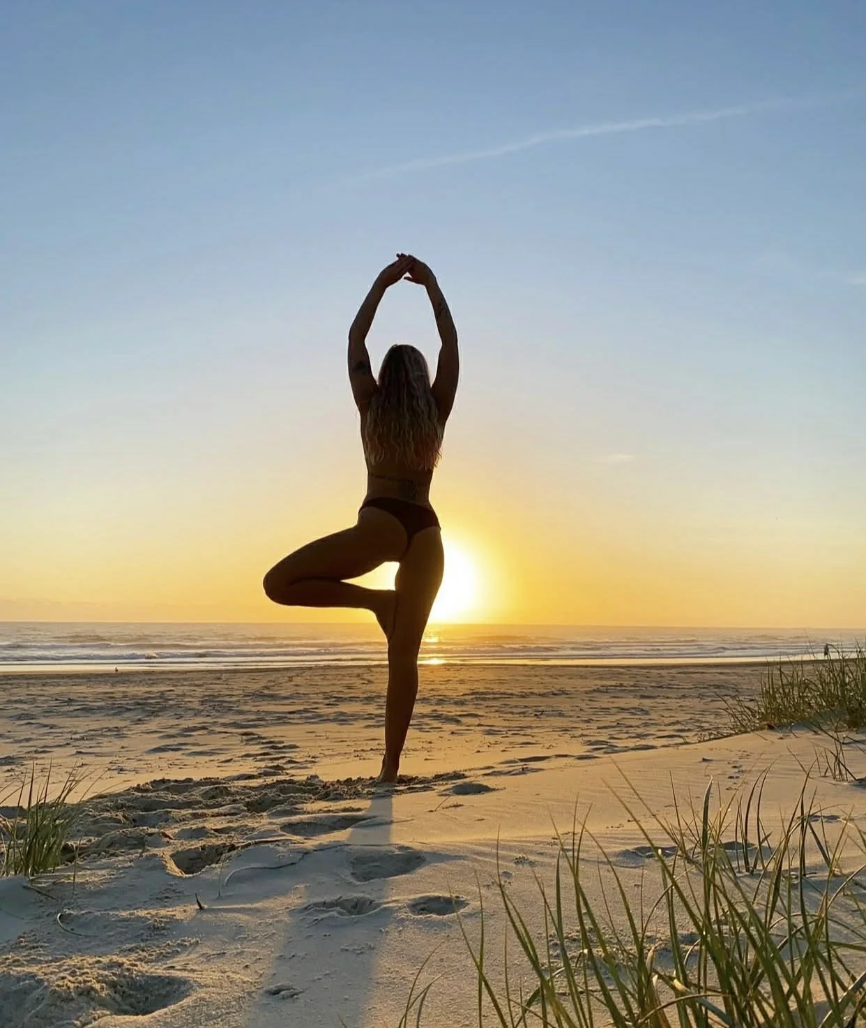 Silhouette of a woman practicing yoga on the beach at sunset, balancing in tree pose with one leg bent, arms raised overhead, and the sun setting over the ocean in the background.