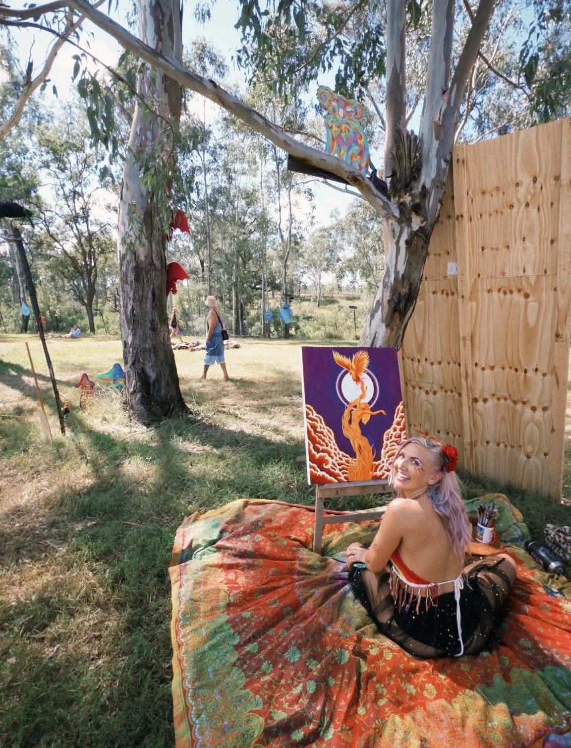 A woman with pink hair and colorful makeup sits on a multicolored blanket in a park, smiling at the camera. She is near an easel with a vibrant fantasy-style painting of a phoenix and a large tree with a swirl of clouds in the background. She is outdoors under trees, with other people visible in the background, and there are various art supplies and decorations around her.