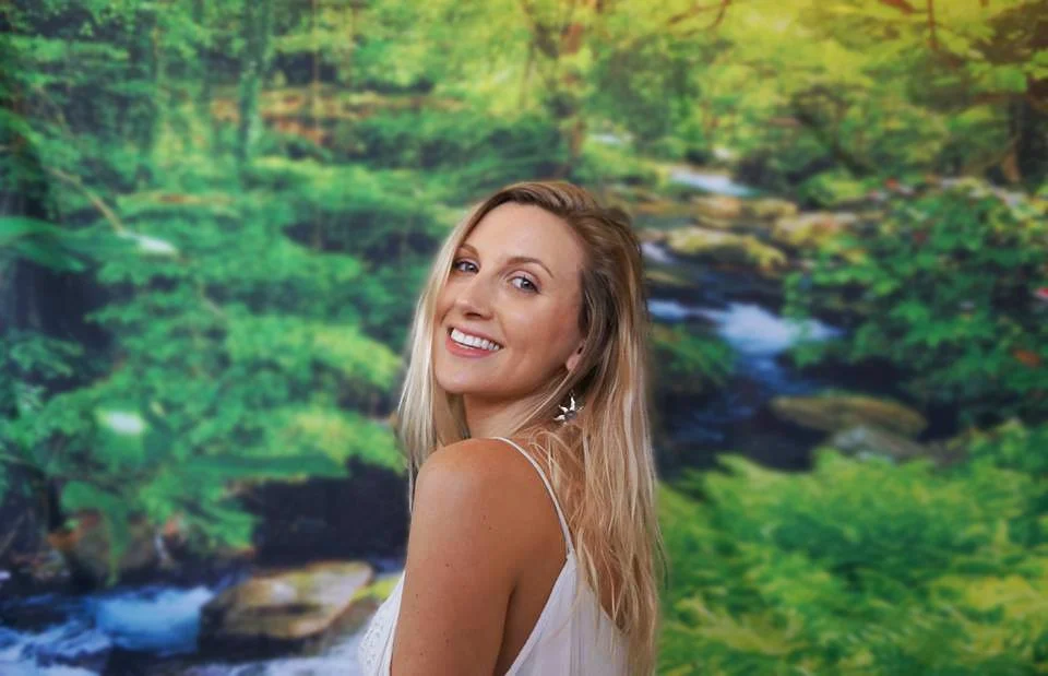 A woman with blonde hair smiling, wearing a white sleeveless top, standing in front of a green forest background with a stream.