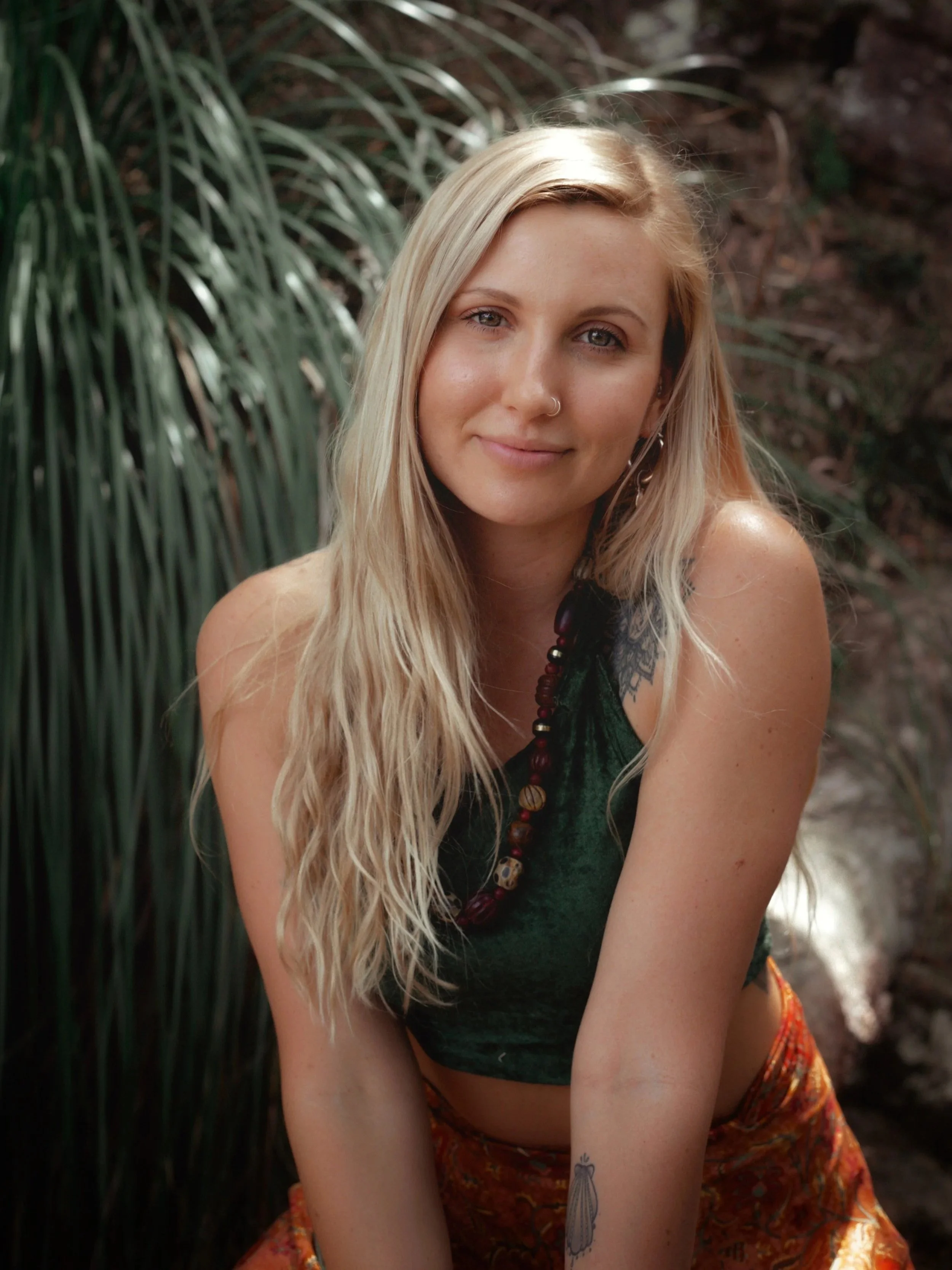 A young woman with long blonde hair, blue eyes, and a nose piercing, sitting outdoors near green plants, wearing a green top, colorful skirt, and a bead necklace.