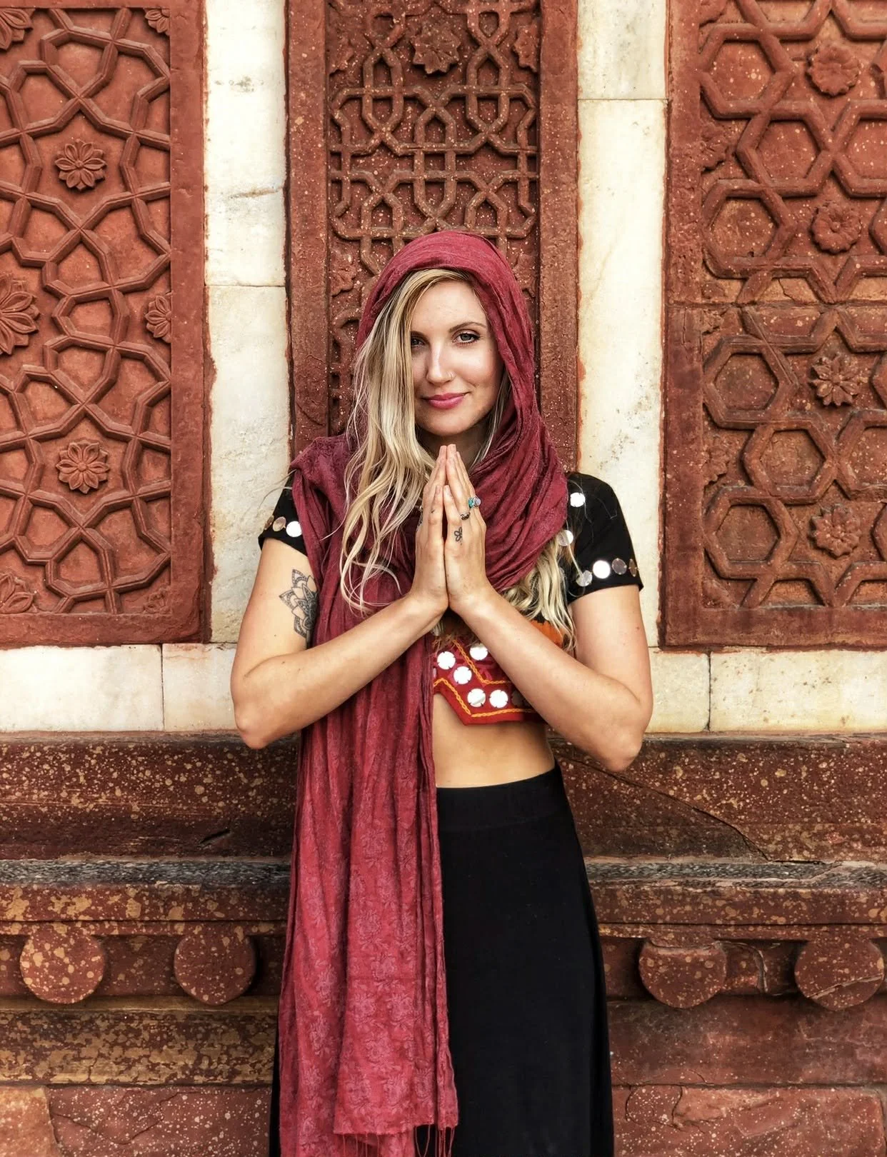 A woman with long blonde hair and tattoos, wearing a black top with white circular decorations and a red scarf, standing in front of an ornate reddish-brown stone wall with geometric carvings. She has her hands pressed together in a prayer-like gesture.