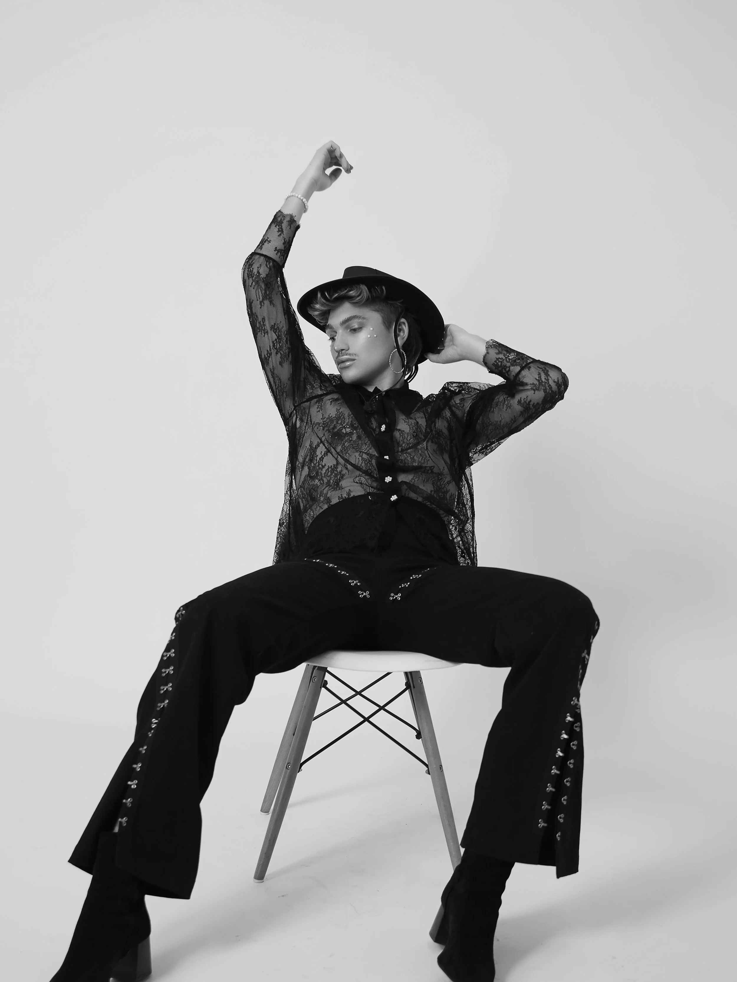 A person sitting on a chair with legs apart, wearing a lace sheer top, wide-leg pants with embellishments, a hat, and high heels, posing with one arm raised and the other behind their head in a studio setting.