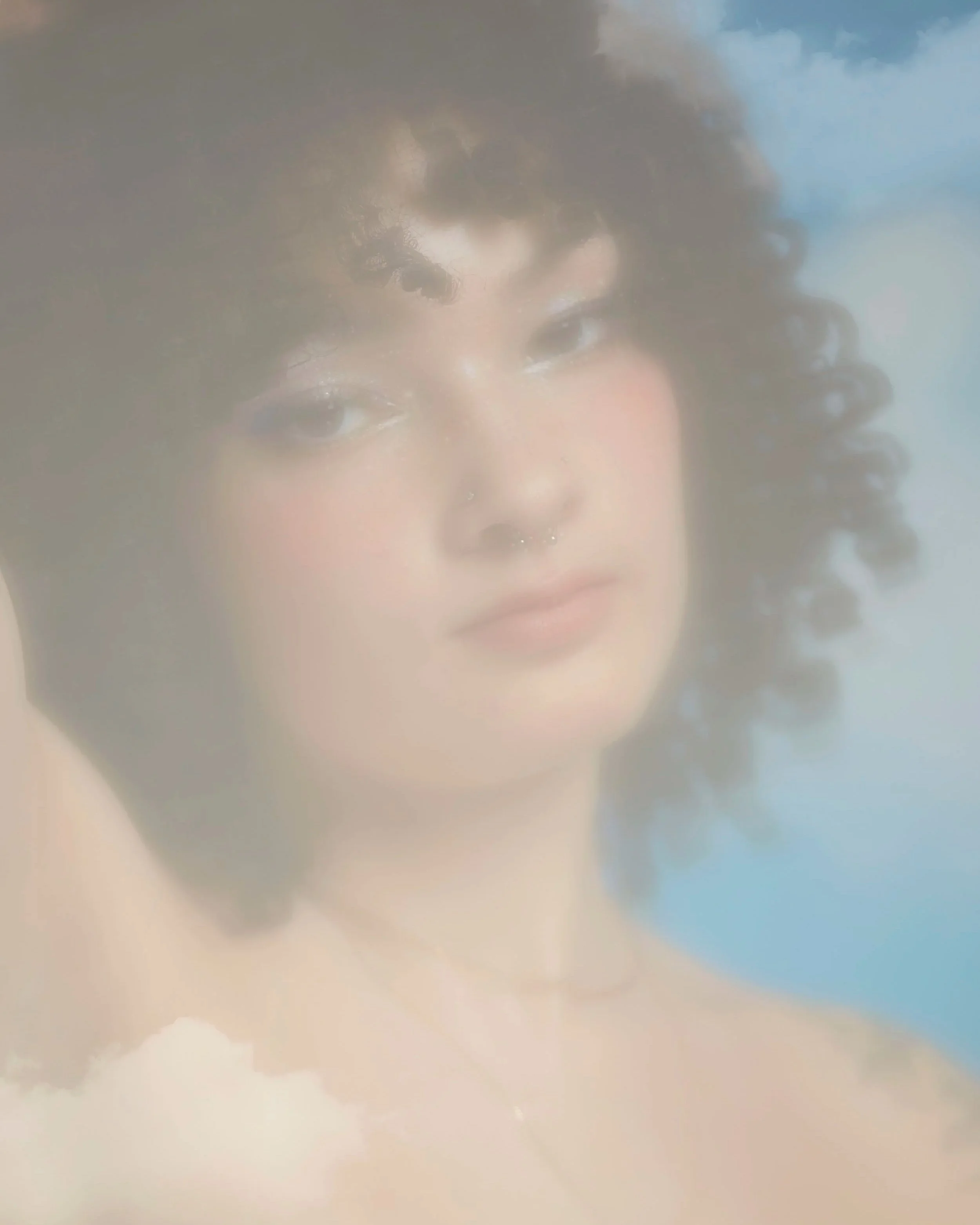 A woman with curly hair, wearing makeup, and a nose ring, looking serene, with a soft focus and sky background.