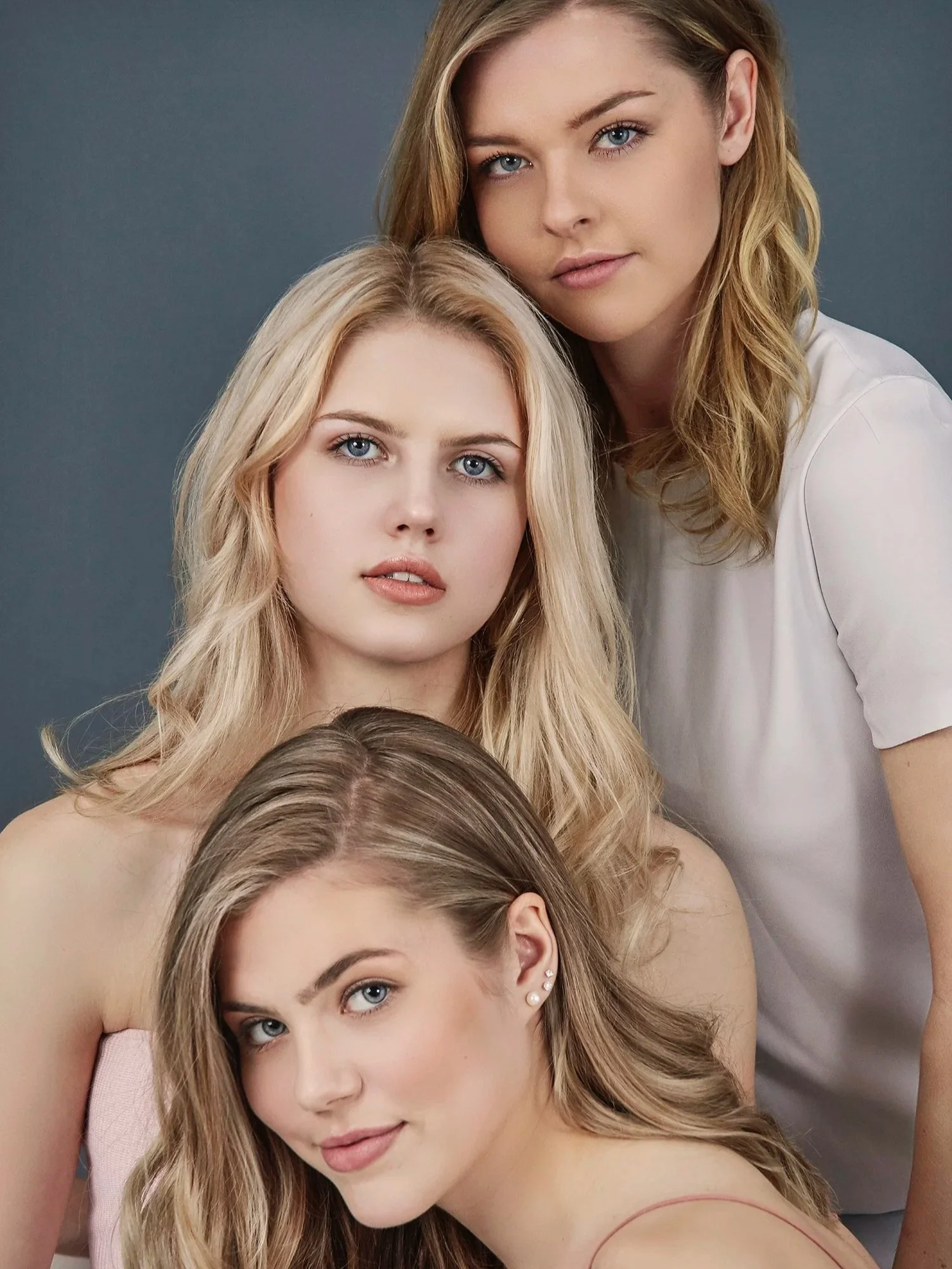 Three young women with blonde hair and blue eyes posing together against a gray background, all looking at the camera.
