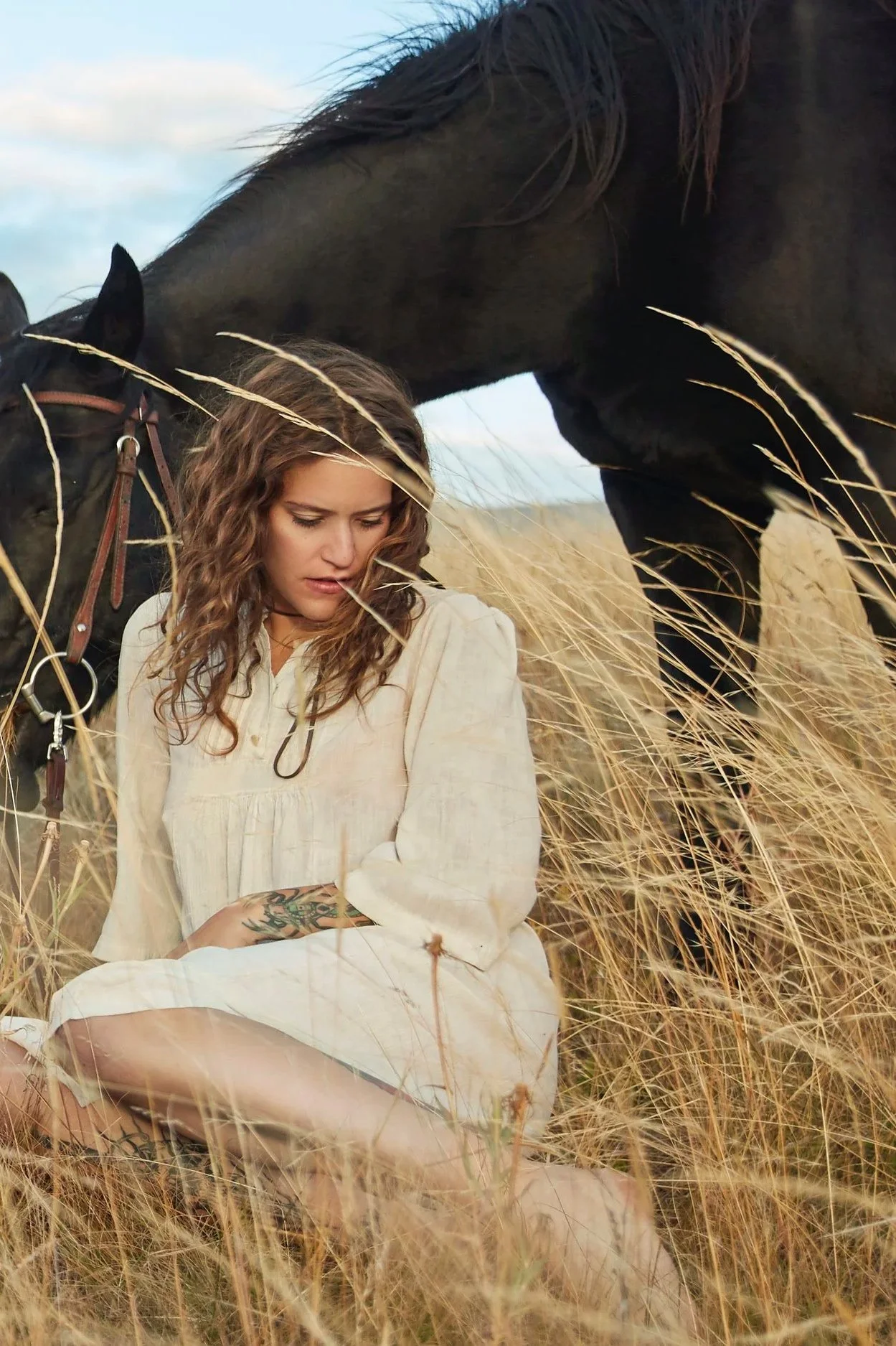 A woman with curly hair and a tattoo on her arm sitting in a field of tall, dry grass next to a black horse. The woman is looking down, and the horse is leaning its head over her. The sky is partly cloudy in the background.