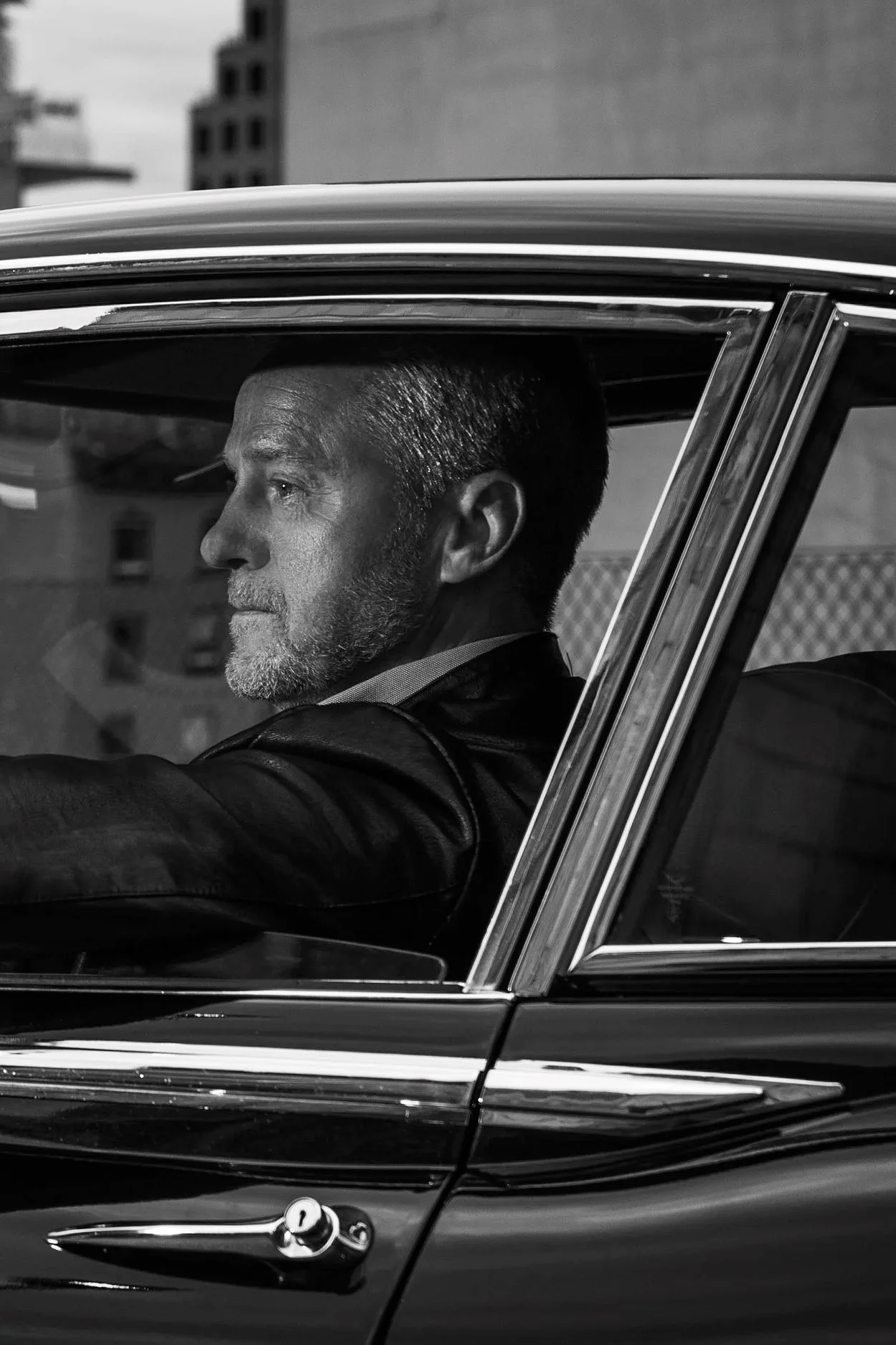 A middle-aged man with short hair and a beard driving a vintage car, seen from the side through the open window, in black and white.
