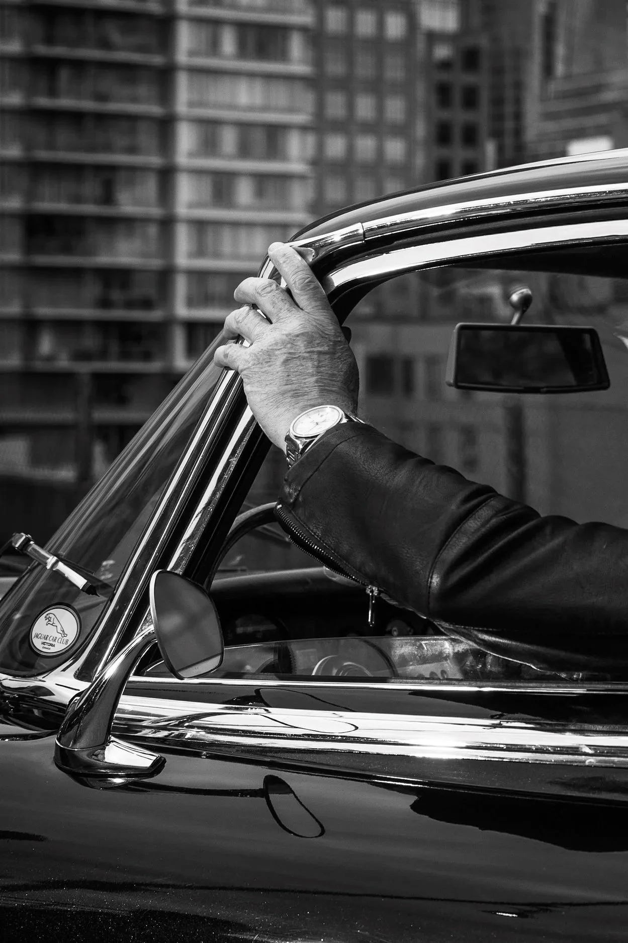 Close-up of a person wearing a watch and leather jacket holding the steering wheel of a vintage car with a cityscape in the background.