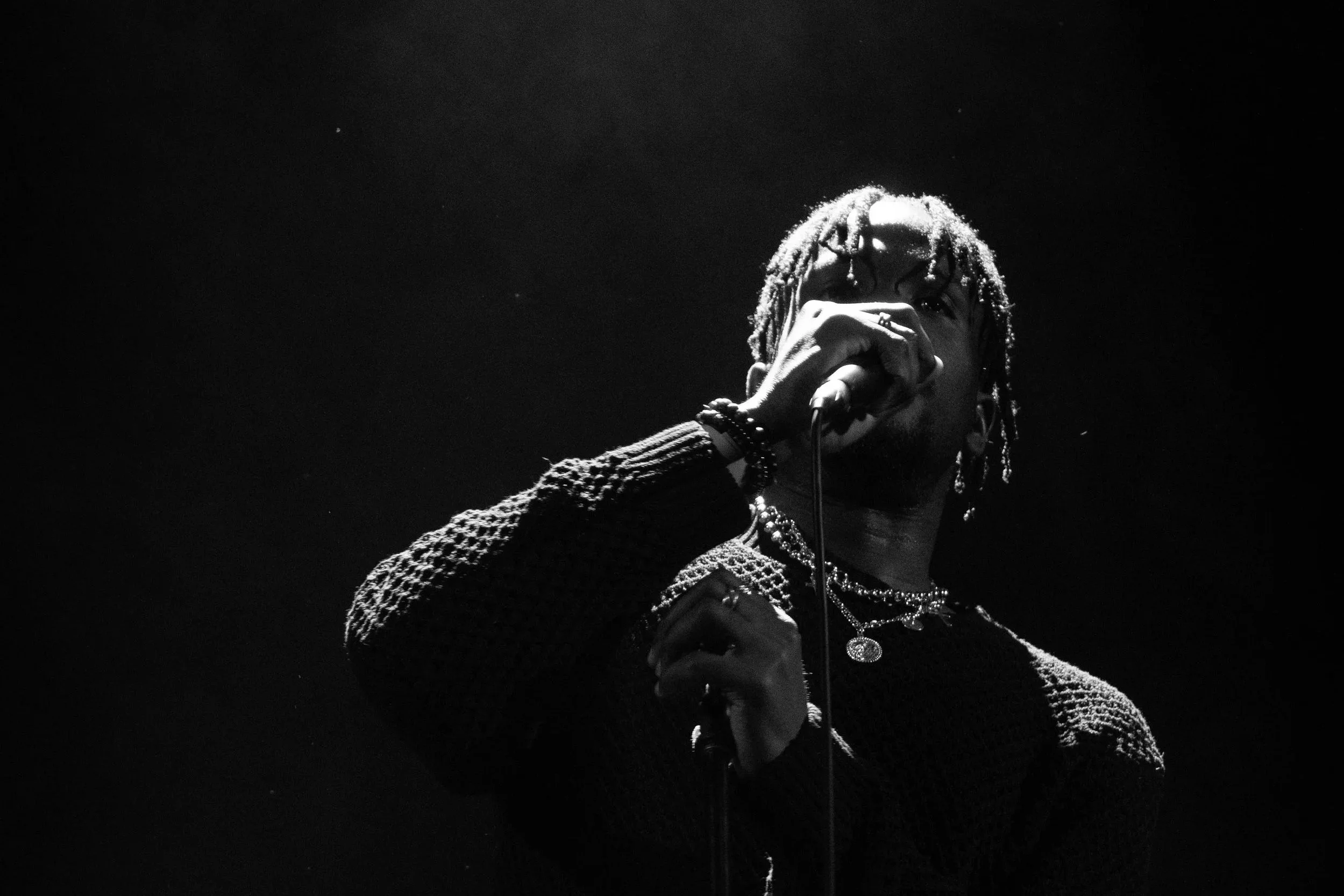 A male singer with dreadlocks singing into a microphone on stage, illuminated by dramatic lighting in black and white.