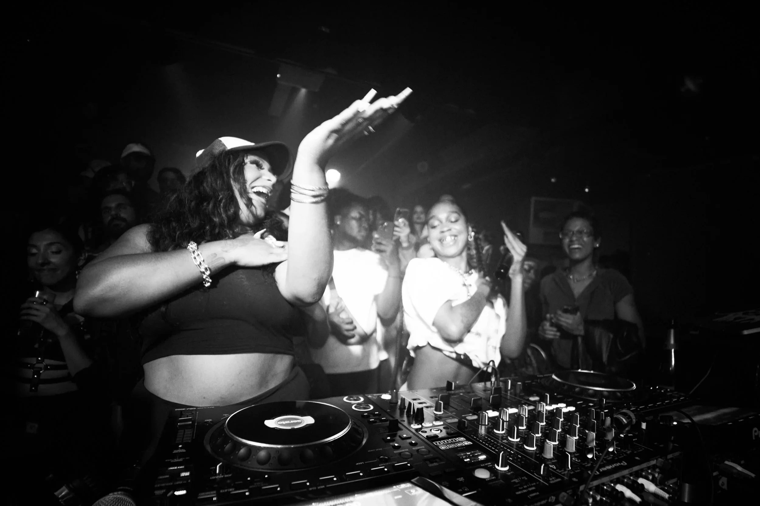 A woman DJ wearing a cap and crop top, smiling and dancing behind DJ equipment, with a crowd of people enjoying her performance.