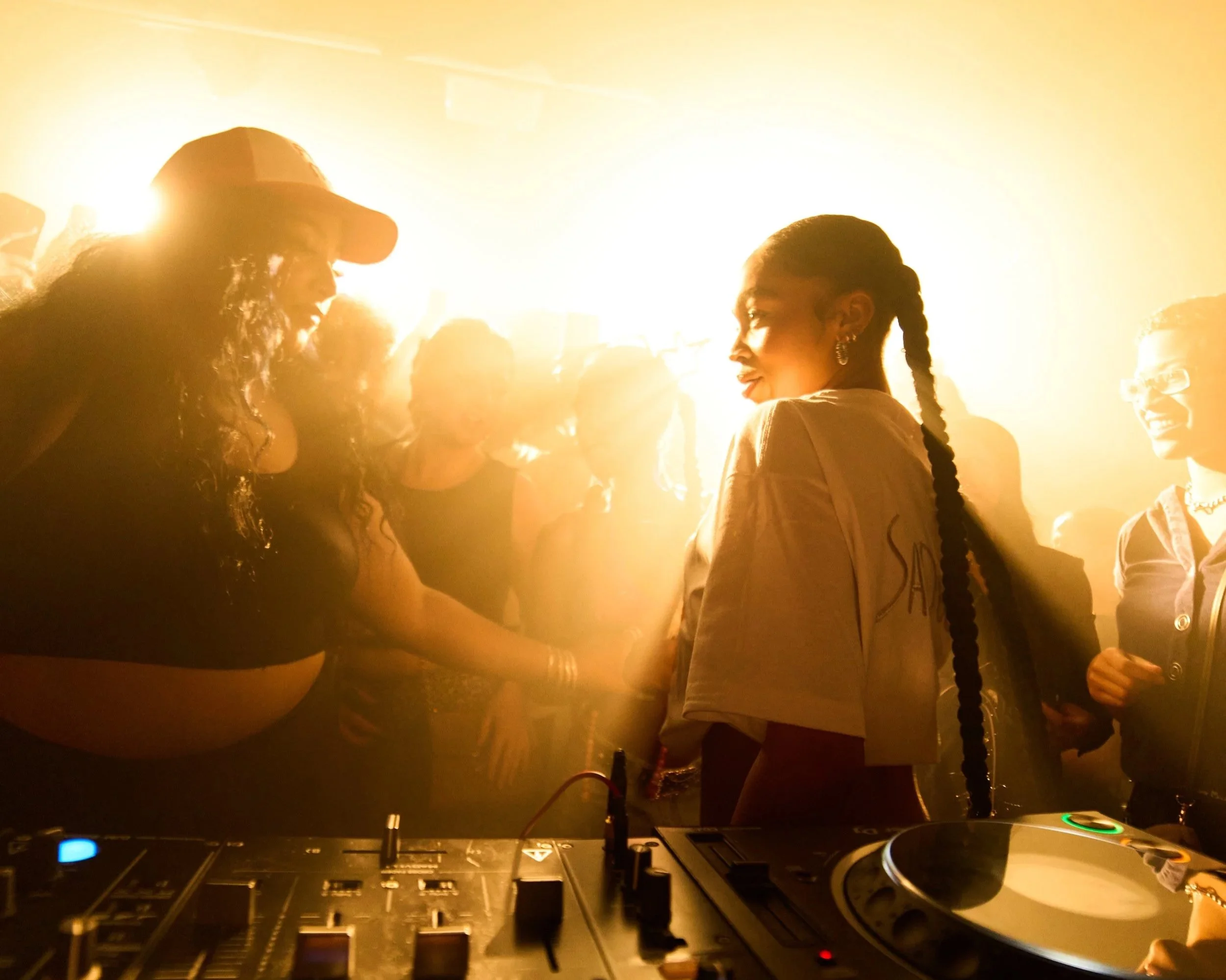 People dancing at a party with a DJ setup in the foreground, illuminated by warm, yellow lighting.