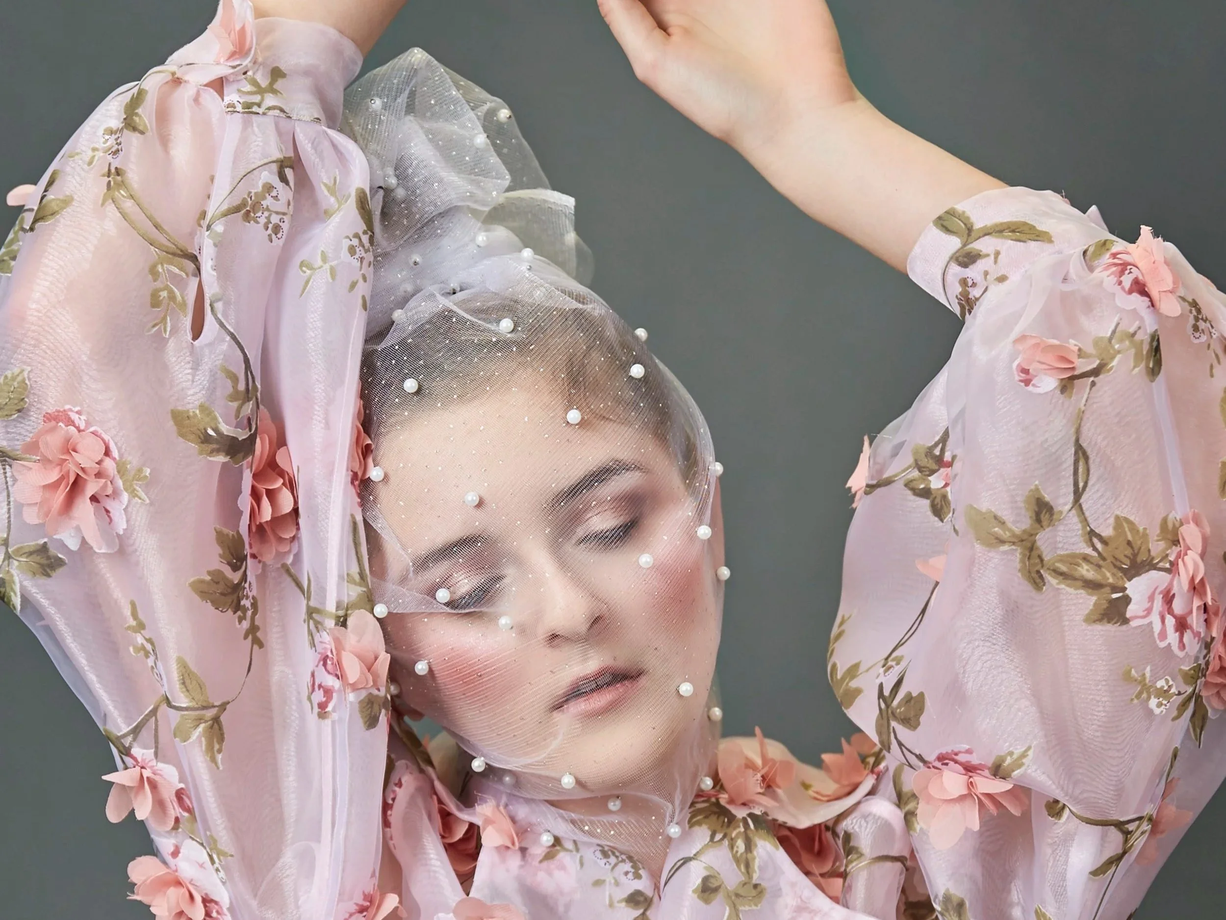 A woman with closed eyes wearing a pink floral dress with puffed sleeves, a sheer veil with pearls, and a matching pink floral headpiece.