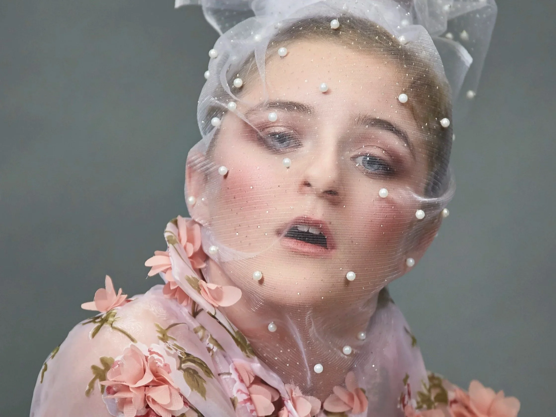A woman wearing a sheer fabric with pearl embellishments over her face, dressed in a floral pink outfit with large pink flowers, has makeup with neutral tones, and blue eyes, creating an artistic, high fashion portrait.